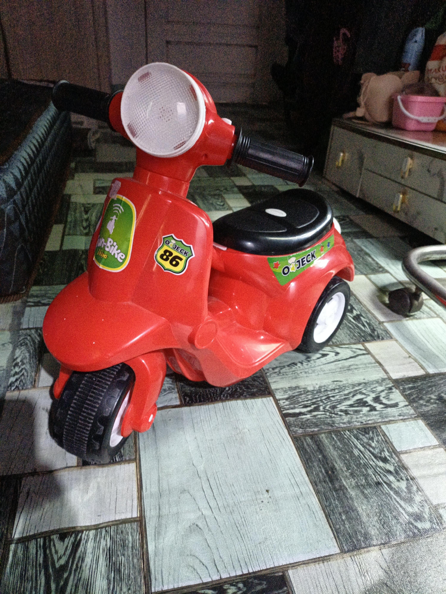 A bright red toy scooter is placed on a patterned floor. The scooter has black handlebars and a white headlight. There are stickers on the scooter: one green sticker with a helmet and the text "Fun-Bike" and "Thailan", and another shield-shaped sticker with "OJECT" and the number "86". The scooter has black wheels with white rims and a black seat. In the background, a dark dresser and some laundry are visible. The floor appears to be made of tiles with a wood-like pattern. The overall scene suggests an indoor setting, possibly a room in a home. The location is identified as Teumpokteungku, Indonesia.