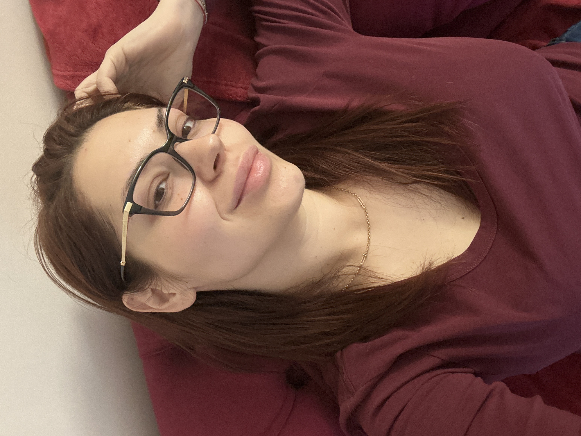 A woman is lying on a red pillow, likely indoors. She is wearing a maroon long-sleeved shirt and glasses with black frames. A delicate gold necklace is visible around her neck. She is looking at the camera with a slight smile. There is no indication of the location being Sofia, Bulgaria in the image.