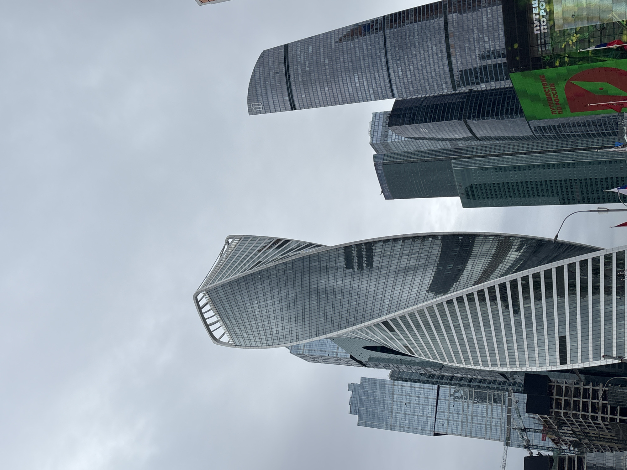 The image shows a low-angle view of several skyscrapers in Moscow, Russia. Prominently featured is the Evolution Tower, characterized by its spiraling design. Other skyscrapers are visible, including one with a large advertisement in Russian. The sky is overcast. Construction work is underway near the base of one of the buildings.