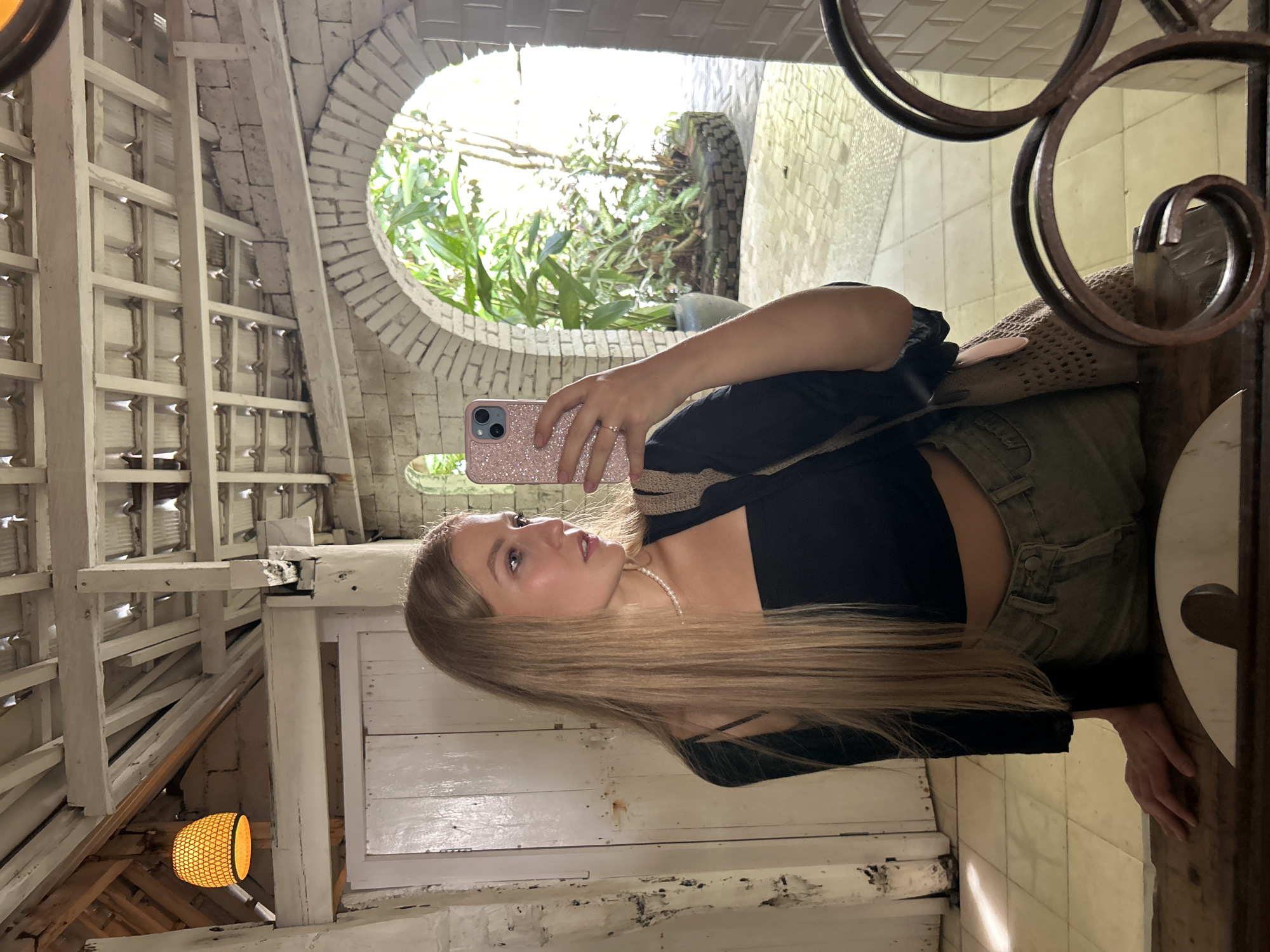 A young woman is captured in a mirror selfie, standing in a rustic, naturally lit interior space. She has long, straight blonde hair and is wearing a black crop top, light-colored high-waisted pants, and a pearl necklace. She holds a pink, glittery smartphone with her right hand, angled to take the photo, while her left hand rests on a surface to her right. Her gaze is directed slightly upwards and to her left, looking into the phone's camera.
The setting features white-washed wooden beams, slats, and a lattice-like ceiling structure. Behind her, a white-painted brick archway opens to an outdoor area filled with lush green foliage and what appears to be part of a vehicle tire. A dark, ornate wrought iron frame or decorative element is visible along the right edge of the mirror. In the lower left corner, an orange, woven light fixture hangs from the ceiling. The floor, visible in parts of the reflection, appears to be made of white tiles.
The bright natural light streaming from the outdoor archway suggests it is daytime. The combination of rustic architecture, tropical greenery, and natural materials aligns with the provided location context of Kuta Selatan, Indonesia. There is no visible text in the image.