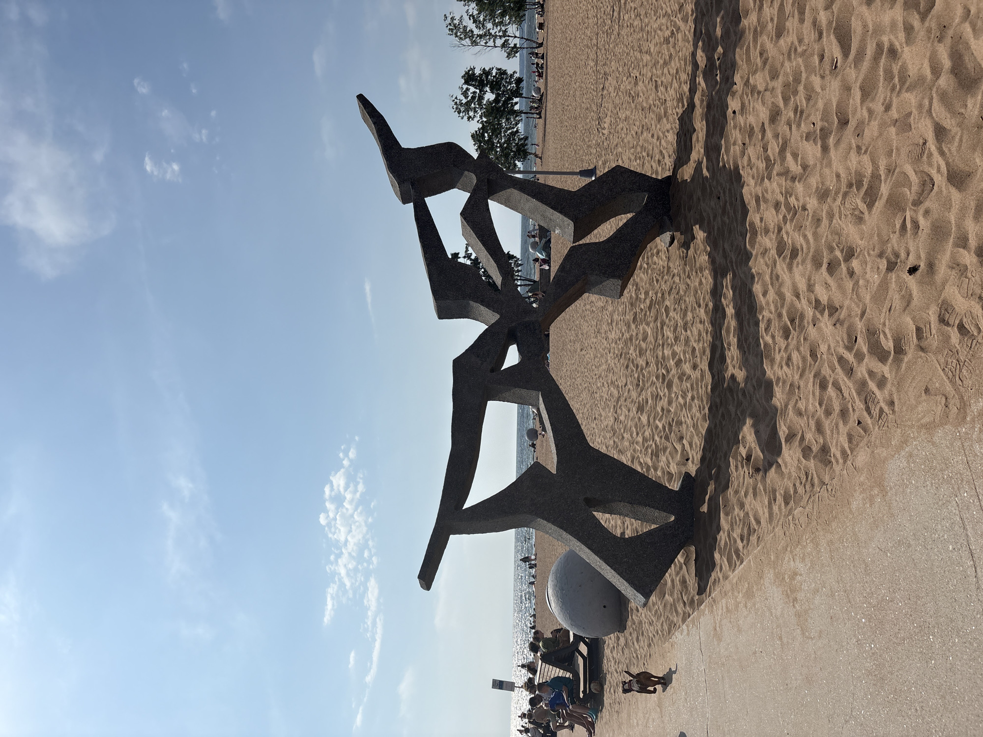 The image depicts a beach scene in Solnechnoye, Russia, under a bright, partly cloudy sky, suggesting a pleasant day, likely in the afternoon given the strong shadows. The foreground is dominated by a large, abstract stone sculpture with sharp angles, casting a prominent shadow on the sandy beach. To the right of the sculpture, a concrete path runs along the edge of the sand.
Beyond the sculpture and path, the sandy beach stretches out, dotted with people relaxing, sitting on benches, and walking. Some are near the water's edge, where the sea is visible, reflecting the sunlight. Trees line the back of the beach, and in the distance, more people can be seen enjoying the seaside. A small dog is visible in the lower right portion of the image, walking on the concrete path. The overall atmosphere appears relaxed and summery, with people enjoying a day at the beach. No visible text is present in the image.