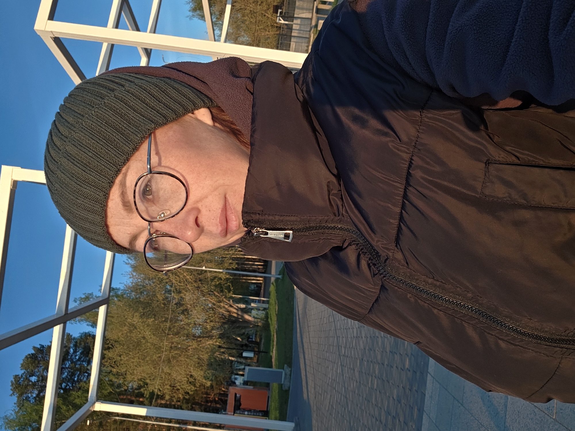 The image shows a person wearing a dark green knitted hat, round glasses, and a black puffer jacket, looking towards the camera. The person is standing on a paved area that appears to be a sidewalk. In the background, there's a modern, white metal structure and trees. Also there are an informational stand and trash cans on the background. The location is the city of Chaikovsky, Russia.
