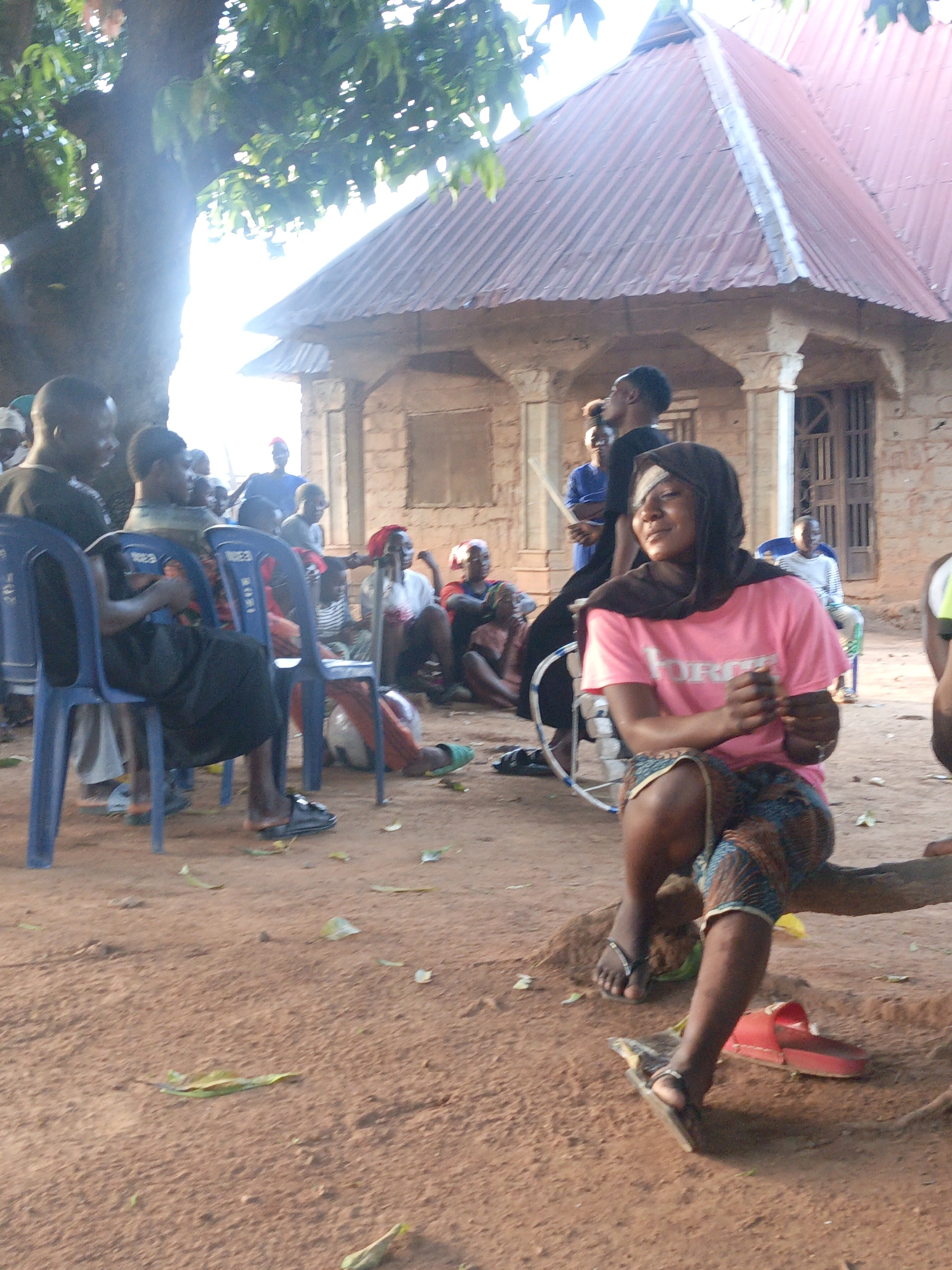 The image shows an outdoor gathering of people in Akwanga, Nigeria. Several individuals are seated on blue plastic chairs, while others sit on the ground or a tree branch. A woman in a pink shirt, patterned skirt, and a headscarf that covers an eye injury is seated in the foreground on a tree branch. A building with a rusty corrugated metal roof stands in the background. The ground is dirt and scattered with leaves. Some people are interacting, while others appear to be observing or waiting. A bicycle is also visible in the background.