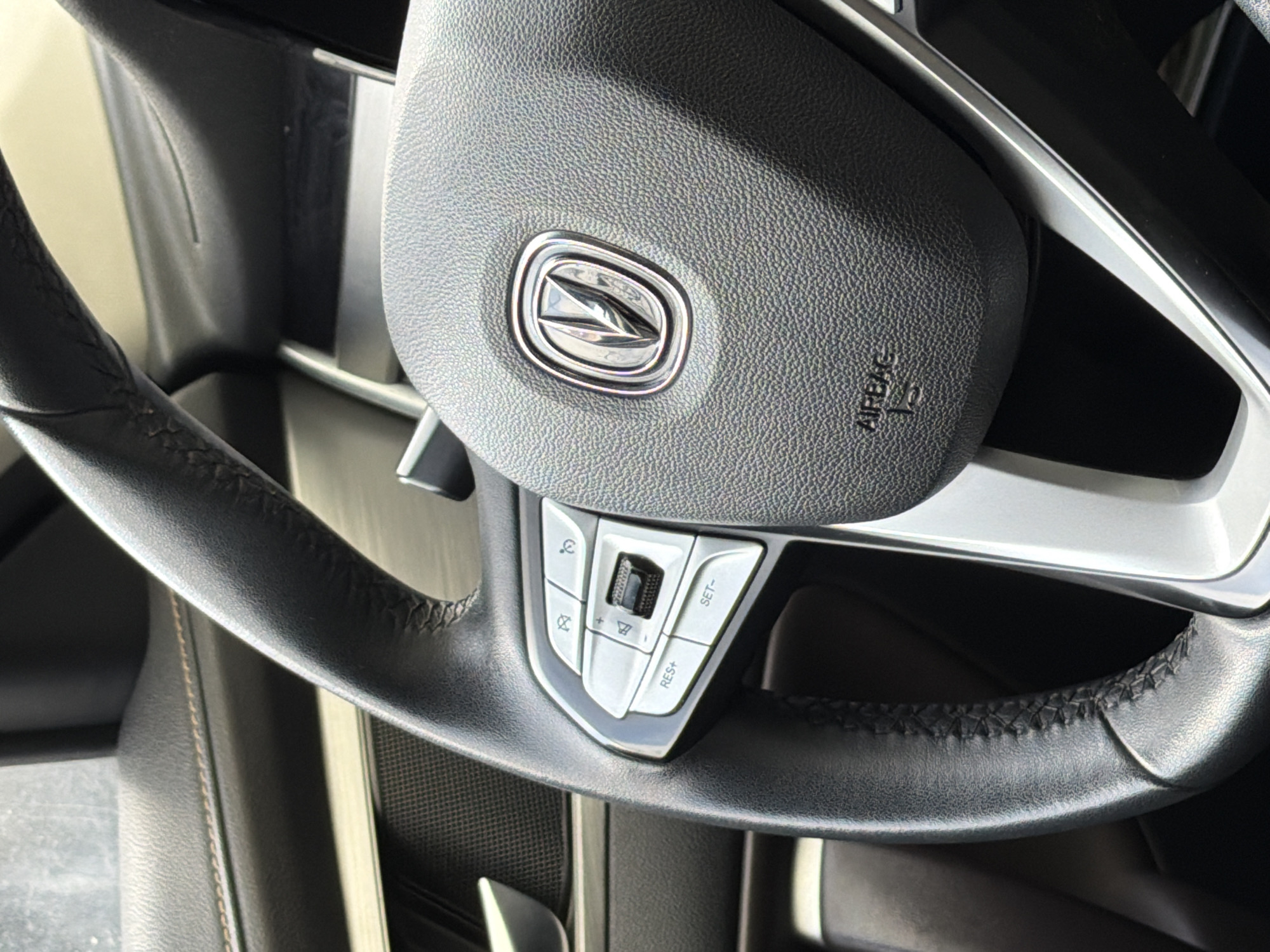 The image shows a close-up of a car's steering wheel. The steering wheel is black and has a silver logo in the center. There are also controls for the radio and cruise control on the steering wheel. The vehicle's make and model cannot be determined from the image. The location is not discernible from the image. No people are visible.
