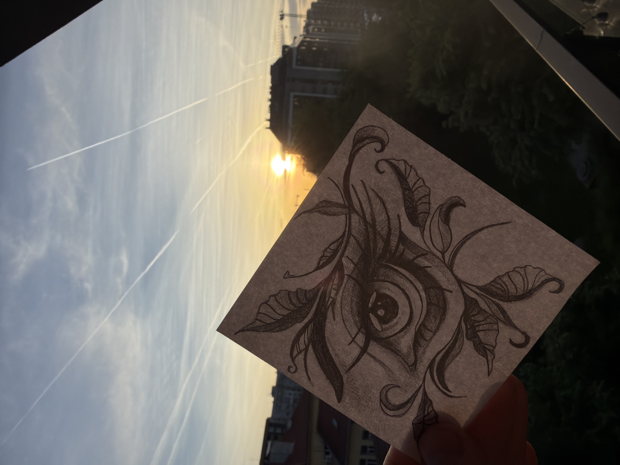 A low-angle, outdoor shot depicts a hand holding a square piece of paper with a detailed pencil drawing of an eye surrounded by leaves and curls, held up against a background cityscape at sunset. The sky is streaked with contrails. A tall building with a construction crane is visible in the background, partially obscured by trees. The photograph is taken from a balcony or window.
