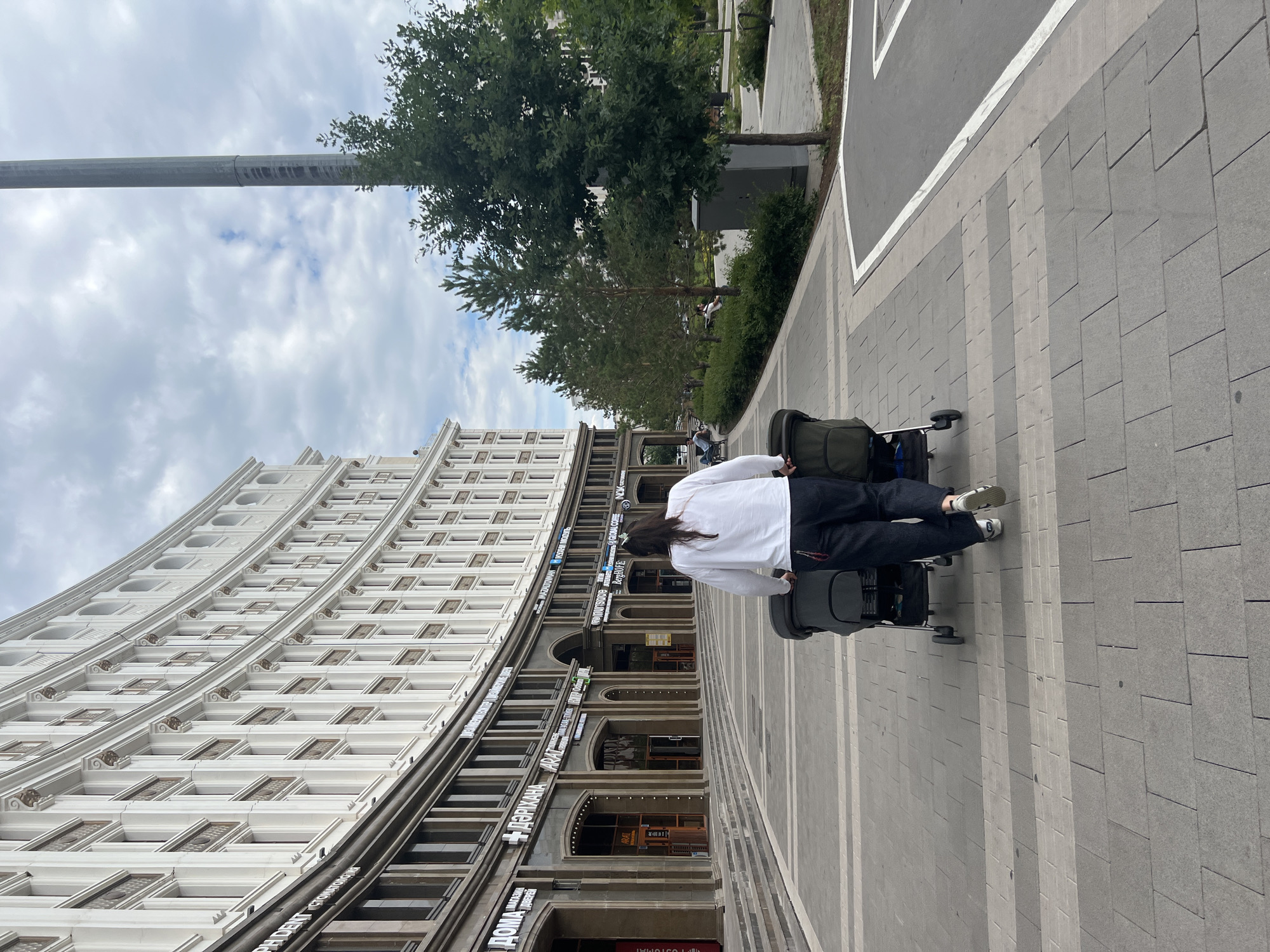 In the city of Astana, Kazakhstan, a person with long dark hair, wearing a white long-sleeved shirt and dark pants, is pushing a double stroller along a wide, modern paved walkway. The walkway is bordered by a building with a white, curved facade featuring numerous windows and decorative elements, and by a green area with trees and bushes. A tall, grey pole stands to the right of the frame. The sky is overcast with scattered clouds. The overall scene depicts an urban environment with contemporary architecture.