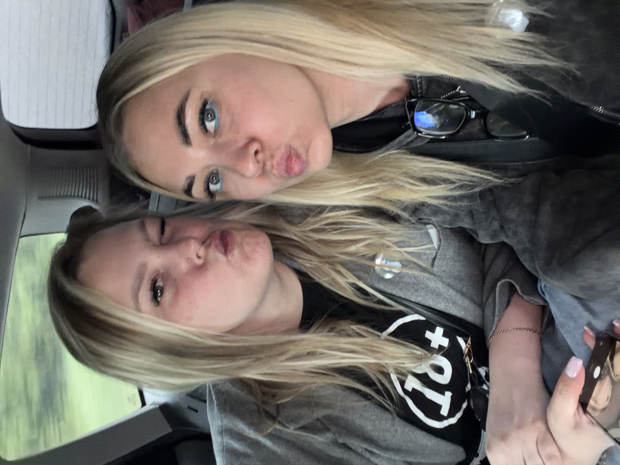 Two young women with blonde hair are in a car, pouting their lips at the camera. The woman on top has blue eyeliner and glasses on the side of her head. The woman on the bottom is wearing a black shirt with a white design. The environment appears to be a car interior. The image does not have enough information to establish it's location to the city of Банное, Russia.
