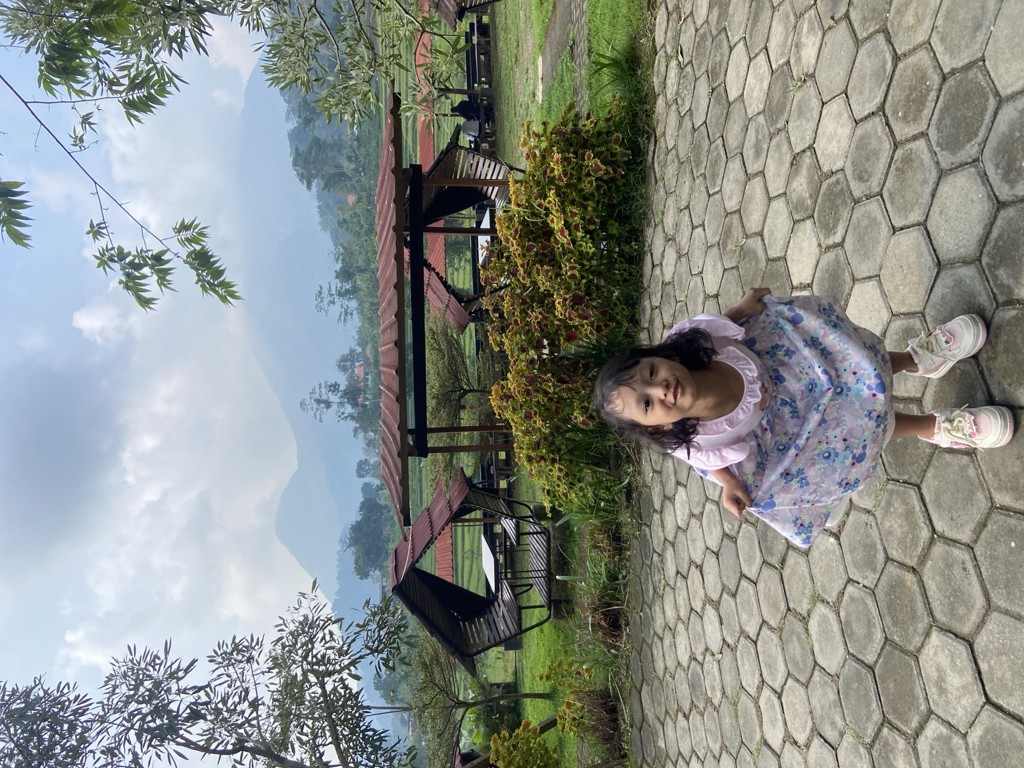 A young girl in a floral dress and pink shoes stands on a gray hexagonal-paved path. Her left hand holds the hem of her dress. In the background, there are open-air pavilions with red roofs and black benches on a green lawn. Lush vegetation, including trees and bushes, surrounds the pavilions. Mountains are visible in the distance under a partly cloudy sky. The location appears to be a park or recreational area, potentially in Penanggungan, Indonesia.