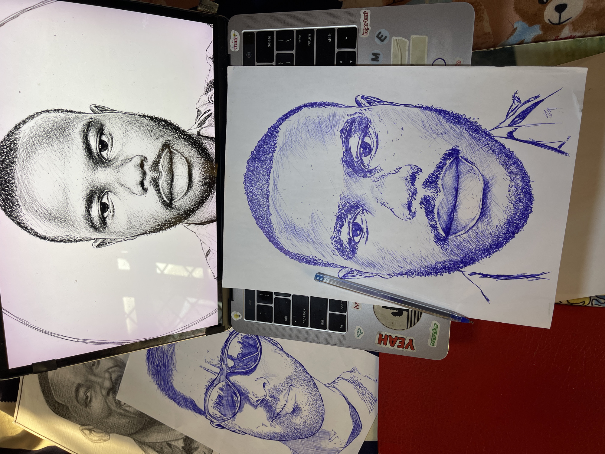The image displays a desk surface with a laptop and several pen-drawn portraits. The laptop is open, showing a digital image of a man's face on its screen, rendered in a black and white sketch style. To the left of the laptop, a physical sketch of a man wearing sunglasses is placed on a red surface. In front of the laptop, another pen-drawn portrait of a man's face is laid out, with a blue pen resting diagonally across it. The sketches are in a detailed, cross-hatching style. The laptop appears to be on a desk with a red surface in the foreground and a textured, possibly fabric, surface visible in the background. Stickers adorn the laptop, including one that reads "YEAH" and another with a cactus illustration. The lighting suggests an indoor environment. There is no information in the image to indicate the location in Jos, Nigeria.