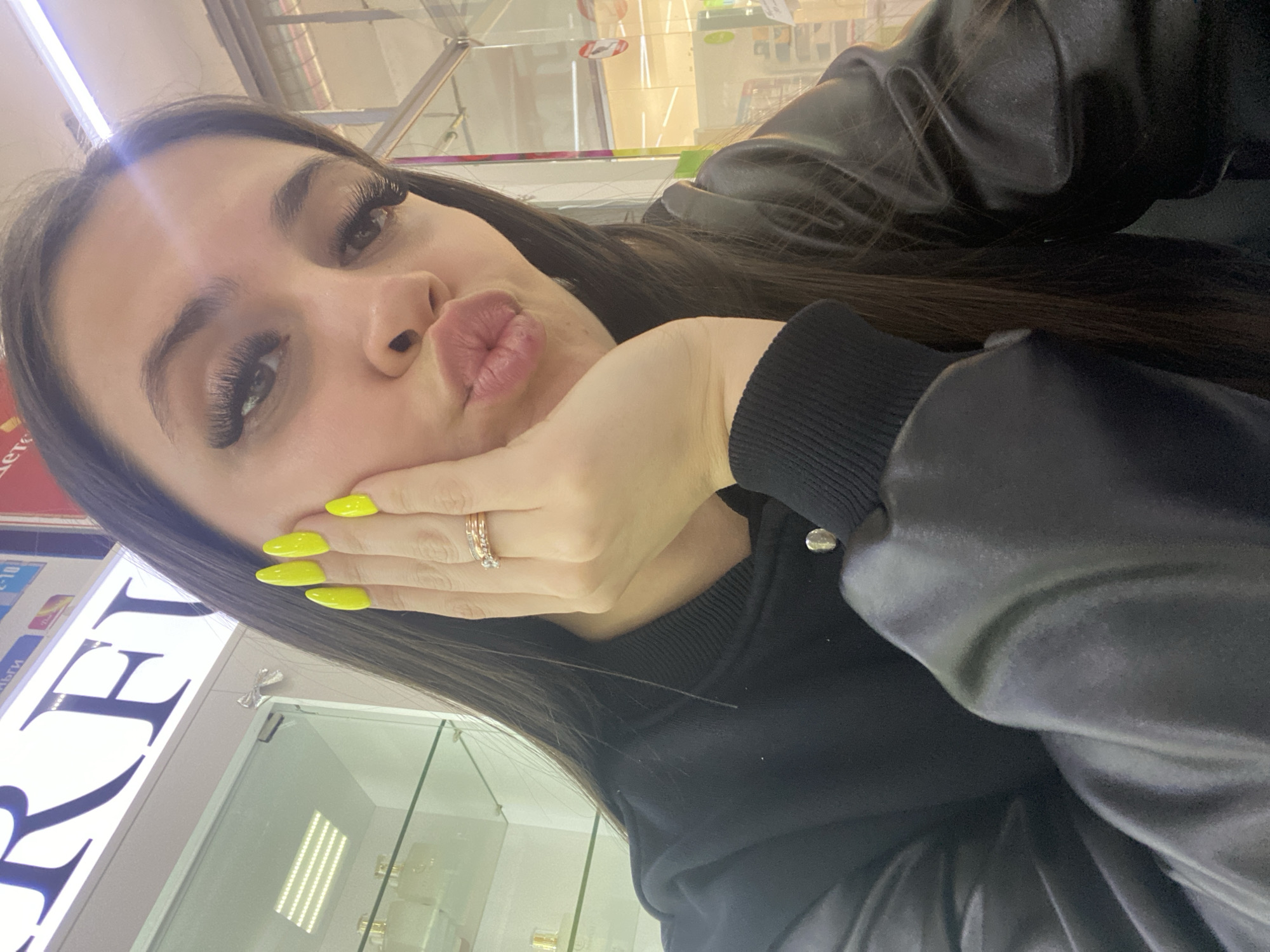 A selfie shows a woman with dark hair, long eyelashes, and bright yellow nail polish. She is pouting her lips and holding her face with her hand, which has a ring on one finger. She wears a black top that appears to be a jacket over a sweater or shirt with a ribbed collar. The background suggests an indoor commercial setting, possibly a shopping mall, with glass display cases and store signage visible. The location is likely Chaikovsky, Russia based on storefront signage.