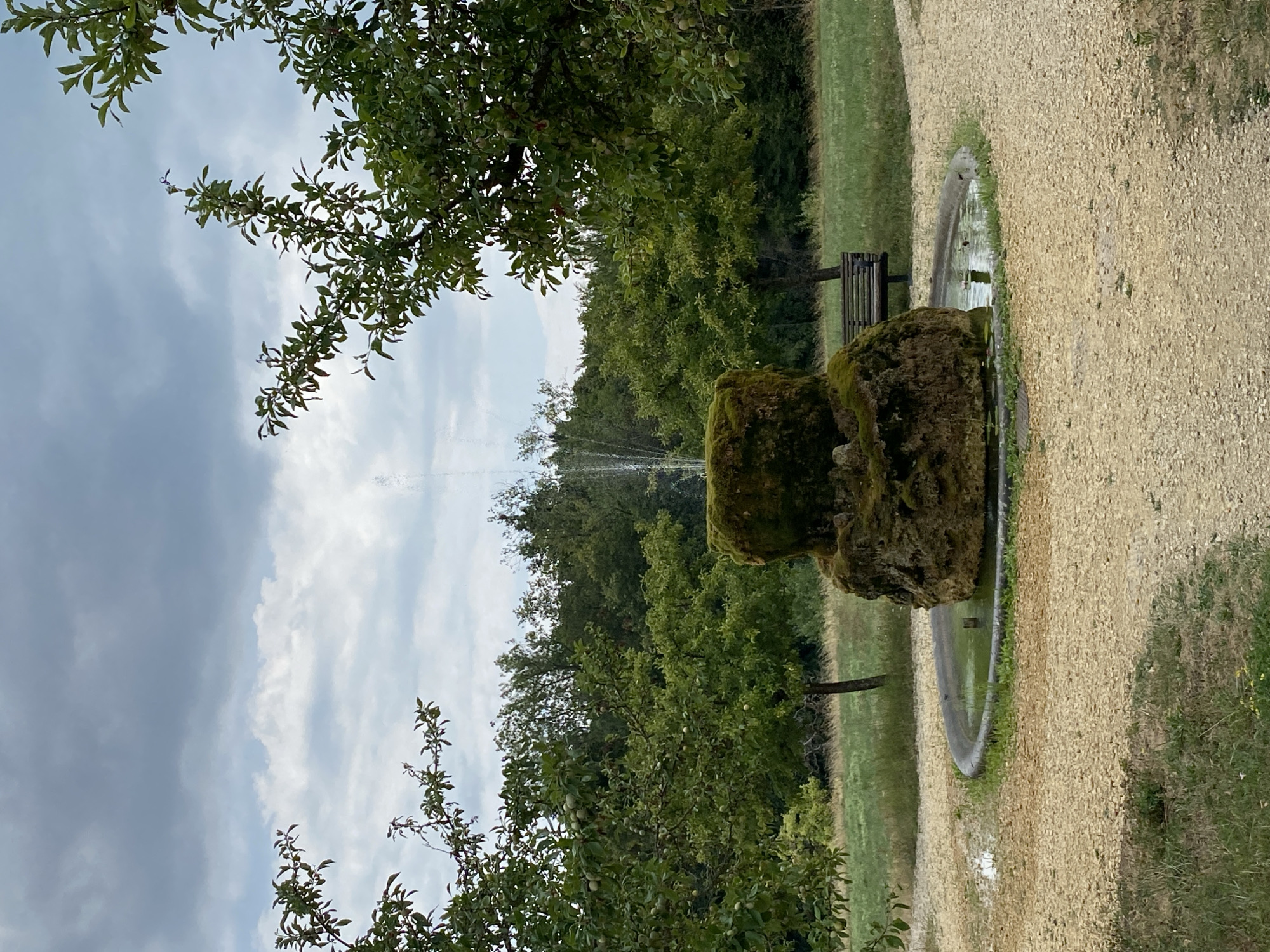 The image depicts an outdoor scene featuring a rustic fountain at its center. The fountain is constructed from rough-hewn stone, covered in moss, and has water spraying upwards. It is situated within a shallow, circular basin filled with green water, surrounded by a gravel pathway. To the right of the fountain, a wooden park bench is visible. The background consists of lush green trees and foliage, with a field stretching beyond. The sky above is overcast with gray clouds. The location is identified as the city of Laténa, Switzerland.
