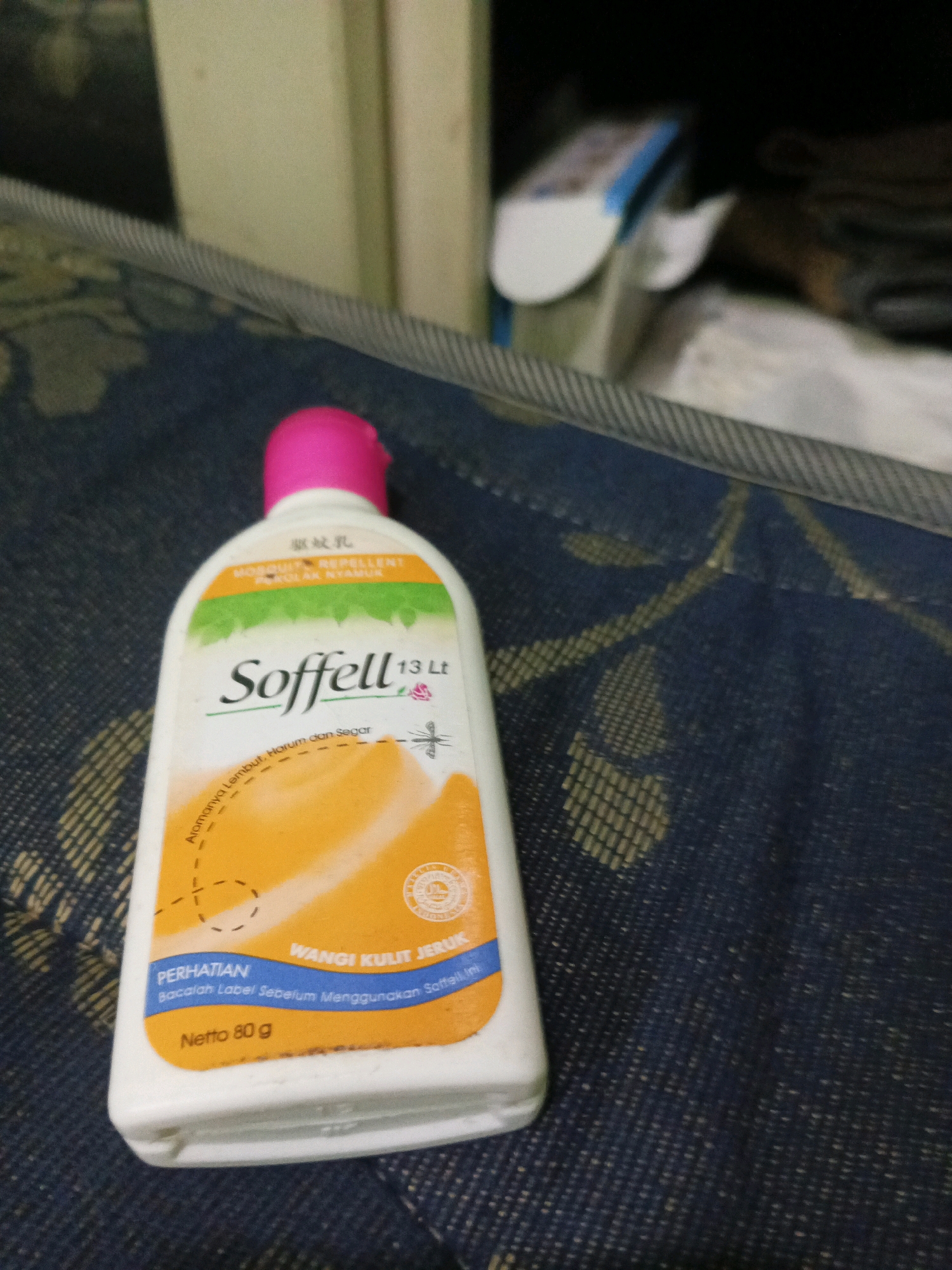 A bottle of Soffell mosquito repellent, with a pink cap, sits on a blue textured surface. The bottle is white and features an orange and green label with text in English and what appears to be Indonesian. The label displays the brand name "Soffell" in large green font, along with "MOSQUITO REPELLENT" and "13 Lt". Below the brand name, there is an illustration of a mosquito and a subtle pattern of orange swirls. There are also sections of text that read "Aromanya Lembut, Harum dan Segar", "PERHATIAN" and "WANGI KULIT JERUK". The bottom of the label indicates "Netto 80 g". The background is out of focus, showing hints of furniture and possibly a wall. The overall impression is that the photograph was taken indoors, likely in a residential setting within the city of South Tangerang, Indonesia, given the product packaging and common use of such items in the region.