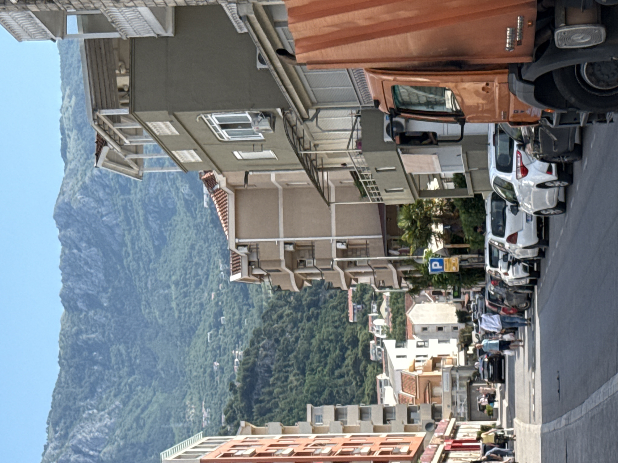 The image depicts a street in Budva, Montenegro, bordered by buildings of varying heights and architectural styles. A large mountain looms in the background on the left, contrasting with the urban environment. On the right side of the street, cars are parked alongside the curb. People are walking on the street, some of whom appear to be tourists. An orange vehicle is parked along the buildings. There is a blue parking sign on the street.