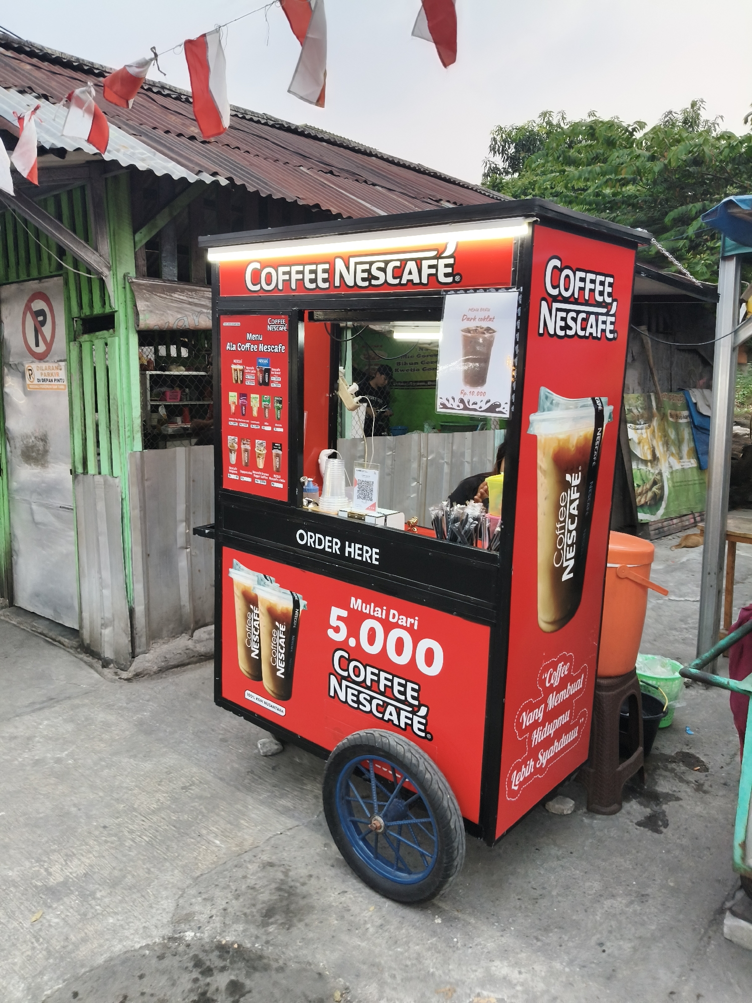 A bright red coffee cart, branded with "COFFEE NESCAFE", is parked on a concrete surface in front of a corrugated metal building with a rusted roof. The cart features a menu board displaying various coffee drinks and prices. Two iced coffee cups with the Nescafe logo are prominently displayed on the side of the cart, alongside text that reads "Mulai Dari 5.000 COFFEE NESCAFE" and "Coffee Yang Membuat Hidupmu Lebih Syahduuu". The cart has a blue-rimmed wheel on its right side. Indonesian flags are strung across the top of the building. A "No Parking" sign is visible on the left side of the image, in front of a green metal gate. The environment appears to be a street or alleyway in a somewhat urban setting, possibly during the daytime given the ambient light. The general appearance suggests a street vendor operation in South Tangerang, Indonesia.