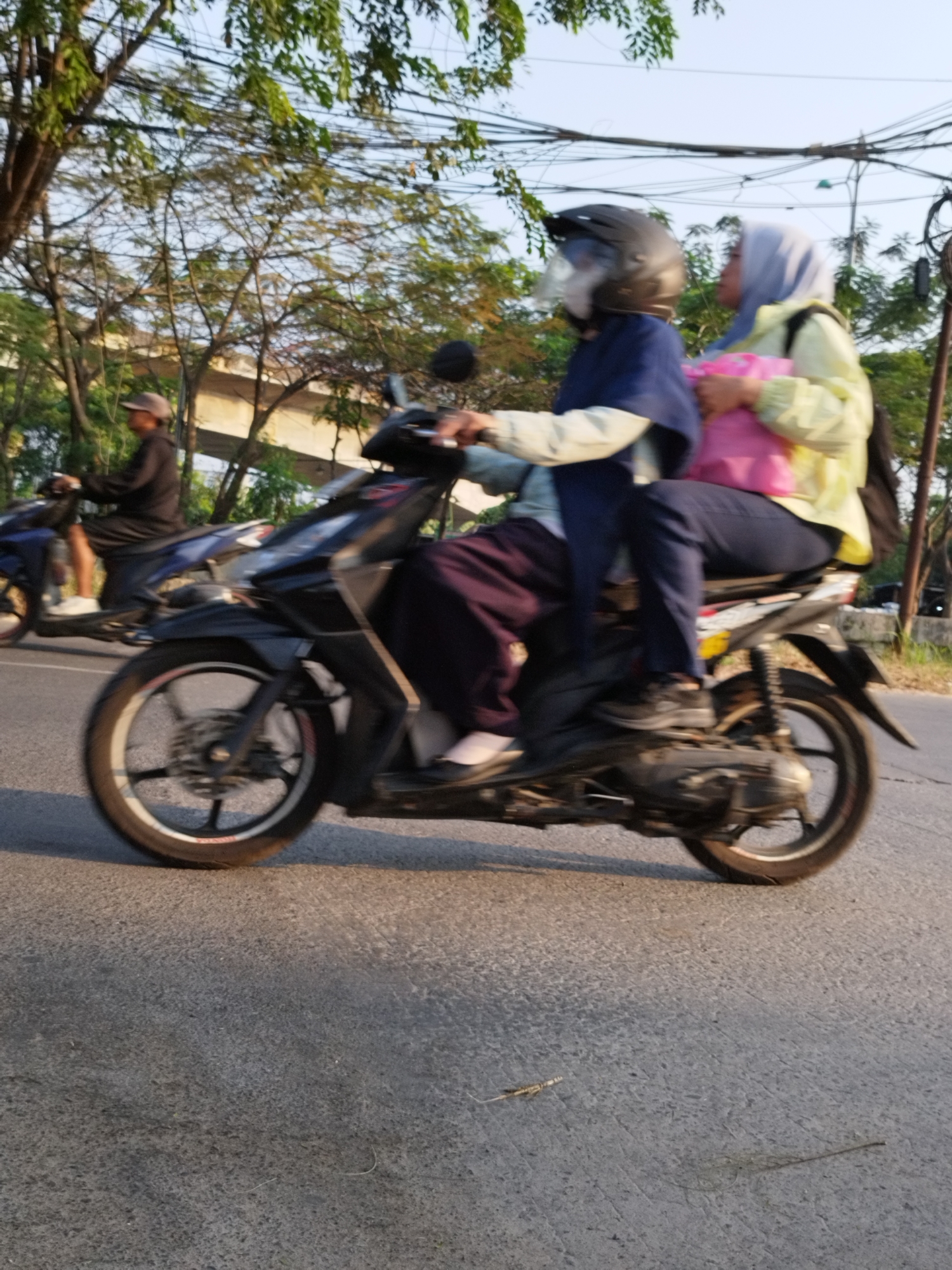Two people are riding on a motorcycle on a paved road. The person in the front is wearing a dark blue headscarf and a light-colored jacket, and is operating the motorcycle. The person in the back is wearing a light blue headscarf and a yellow jacket, and is holding a bright pink bag. Another motorcycle with a person in a dark jacket and a cap is visible in the background, to the left. The scene appears to be in an urban or suburban environment, with trees and power lines visible in the background. The lighting suggests it is daytime.