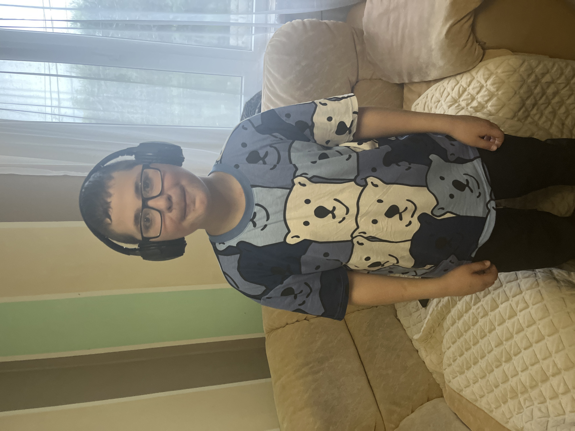 A young boy wearing glasses and headphones sits on a couch in what appears to be an indoor setting. The boy is wearing a blue t-shirt with a pattern of bear faces. He is positioned in front of a window with blinds or curtains. The couch has tan-colored cushions. A multi-colored wall is partially visible on the left. The city and location of Oryol-Izumrud, Russia, are not verifiable within the image.