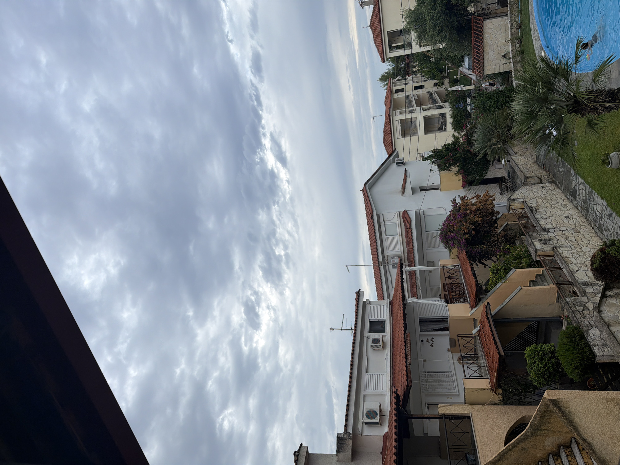The image shows a residential area with multiple buildings and a swimming pool in Pallini Municipal Unit, Greece. The buildings are of various sizes and colors, with red tiled roofs. Some buildings have balconies and air conditioning units visible. There are trees and plants around the buildings, and a stone wall lining a pathway. A bright blue swimming pool is visible in the foreground, with one person swimming in it. The sky is cloudy and overcast.