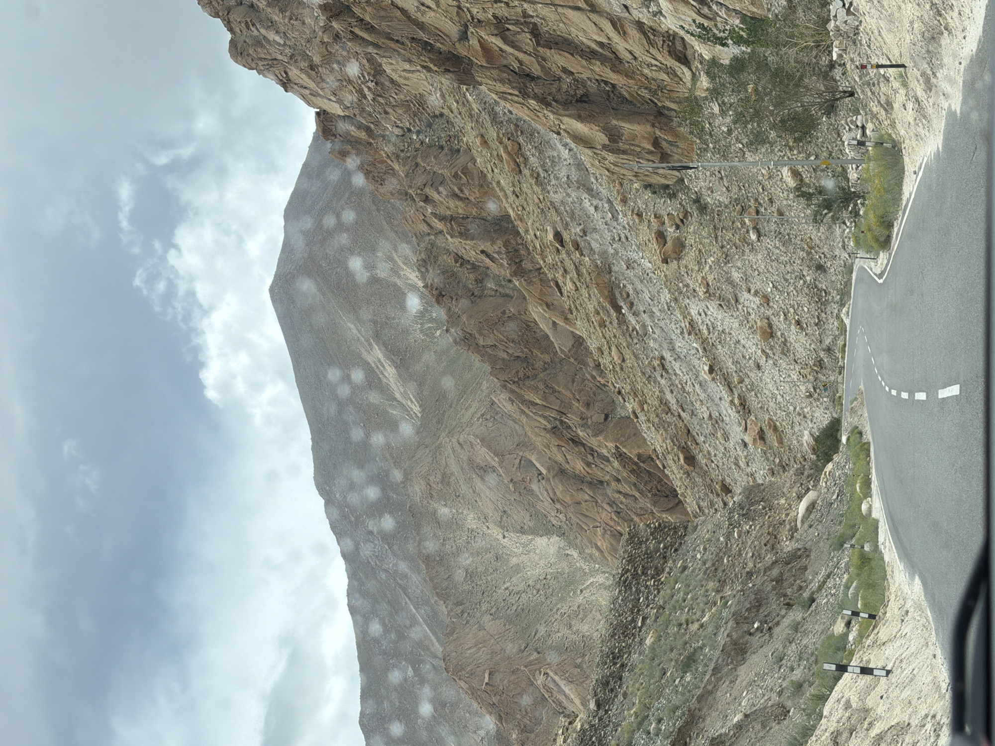 The image depicts a winding asphalt road cutting through a mountainous landscape. The mountains are rugged and barren, with exposed rock faces in shades of brown and gray. Sparse vegetation, consisting of dry grasses and a few small trees, clings to the slopes. The sky is overcast with a pale gray and white cloud cover. Several small, circular, whitish-blue spots are scattered across the image, suggesting water droplets on a lens or window. The road has a white dashed line marking the center. Road markers with black and white stripes are visible on the left side of the road, and a single utility pole stands on the right. The scene appears to be a remote, natural environment, possibly a mountain pass or valley, given the terrain and lack of visible infrastructure. The location is identified as Likche, India.