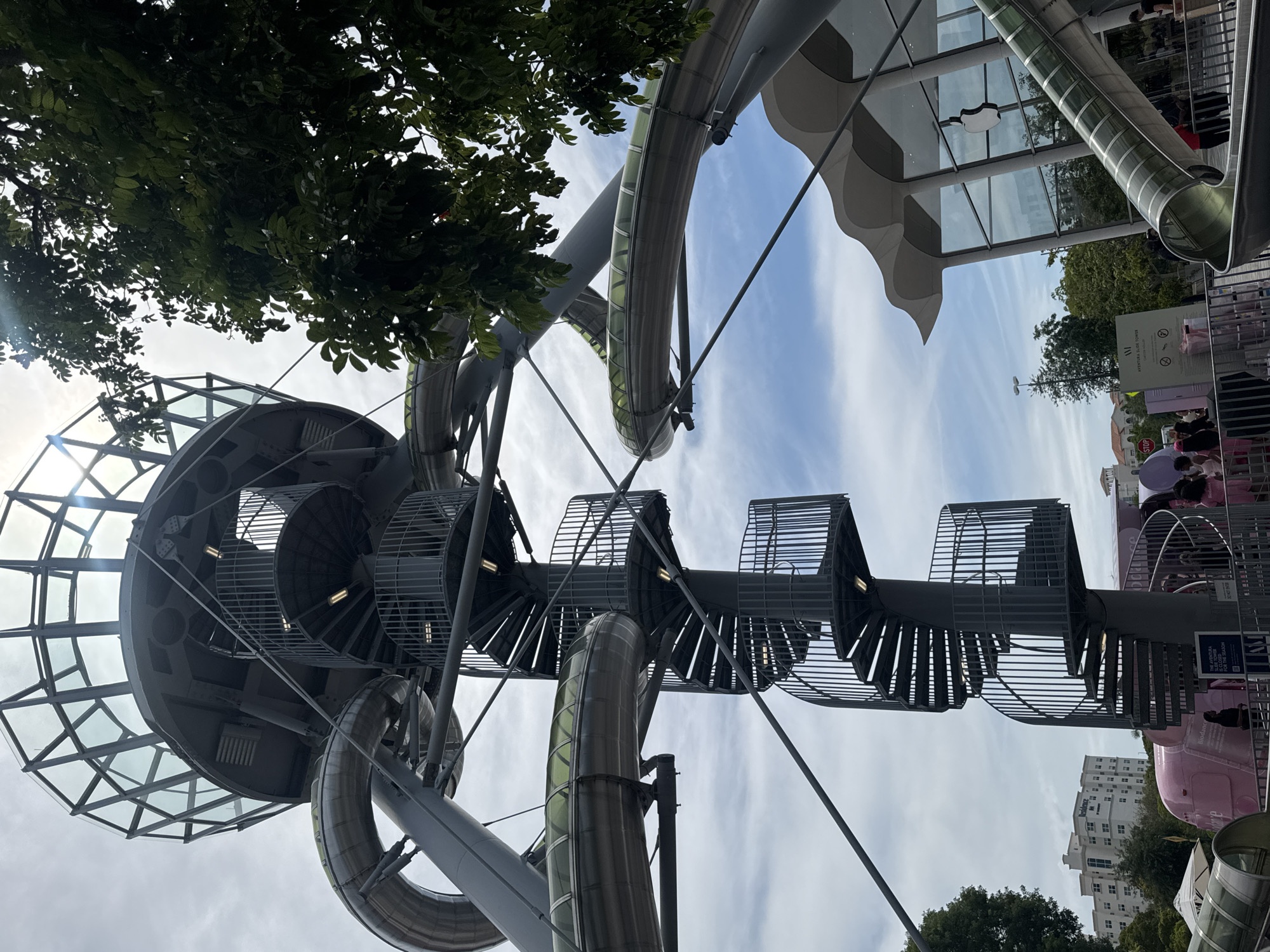 This is an eye-level, exterior, medium shot of a tall, gray, spiral staircase ascending a tower with a glass-domed top. Several large, metal, tube-shaped slides twist and loop around the staircase and the tower. A large, green tree with dense foliage is on the right side of the image, partially obscuring the sky and the top of the tower. In the background, a modern building with a wavy, white roof and large glass windows, featuring an Apple logo, is visible. There are also people on the ground level, near a pink trailer or bus with "BioDance" written on it. A sign near the staircase reads "AVENTURA SLIDE TOWER". The sky is partly cloudy with patches of blue. The overall scene suggests an outdoor attraction or a public space with recreational features.