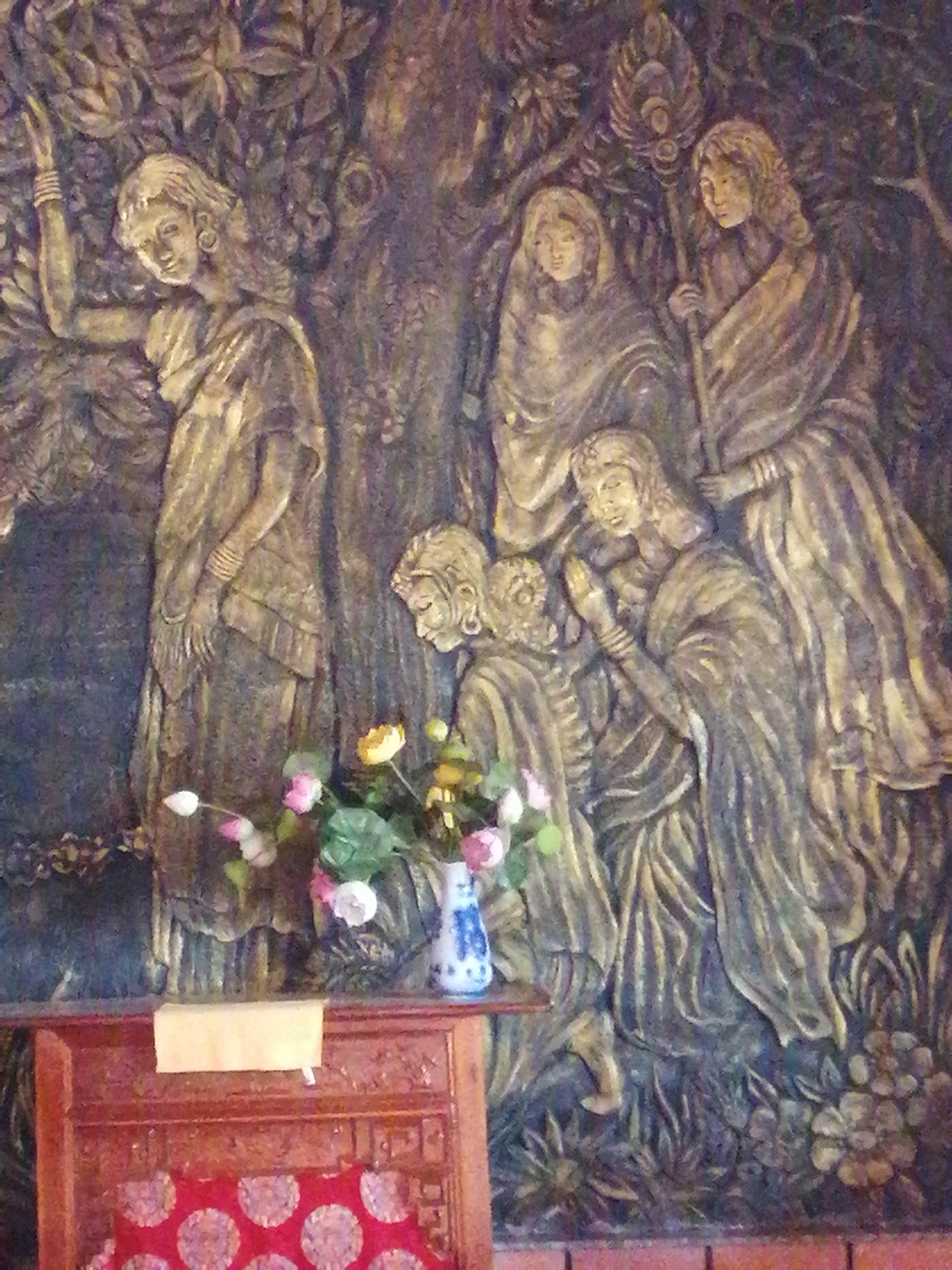 The image is a close-up of a bas-relief sculpture depicting a group of figures in traditional attire, possibly an allegorical or historical scene. The figures are arranged among stylized trees and foliage, rendered in shades of gold and dark brown, giving the impression of aged bronze or wood. The figures, appearing to be women, are adorned with jewelry such as earrings and bracelets, and wear draped garments. Some are standing, while others are seated or bending. In the foreground, artificial flowers in pink and yellow hues are placed in a vase, alongside a wooden stand or altar with carved details and a red, circular-patterned cushion. The overall aesthetic suggests a cultural or religious artwork. The artwork is located at the Tuyen Lam Lake area in Da Lat, Vietnam, likely within the territory of the Truc Lam Zen Monastery.