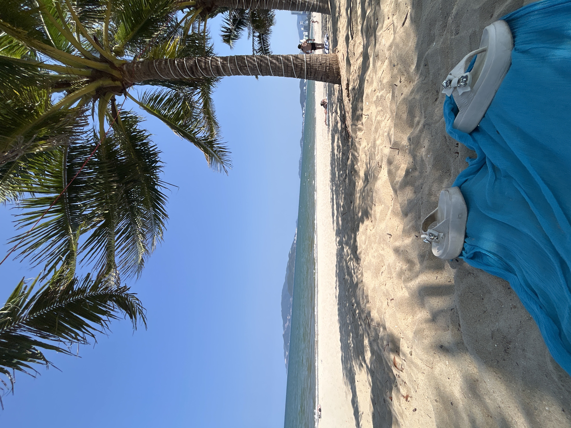 A beach scene in Nha Trang, Vietnam, shows white sand bordered by ocean and a clear blue sky. A palm tree with green fronds leans into the left frame, casting shadows on the sand. A piece of blue fabric sits on the sand with two white sandals with decorative bows resting on top. The beach has several people in the distance. The perspective of the image has been rotated 90 degrees clockwise.