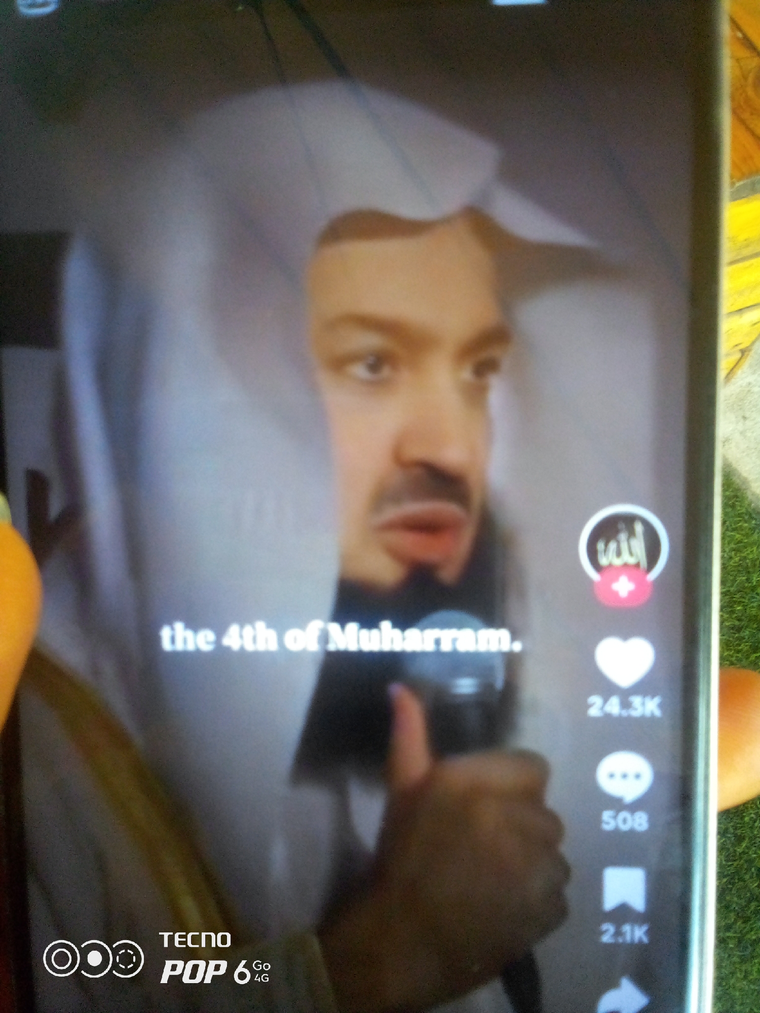 A person is recording a video on a smartphone. The smartphone is displaying a video of a man speaking, wearing a white headscarf. Text overlays on the video read "the 4th of Muharram." The lower portion of the smartphone screen shows the TECNO POP 6 4G logo. The background of the image shows a blurry green surface, possibly grass, and a wooden table. The image appears to be taken outdoors in the city of Howayi, Nigeria, based on the context provided.