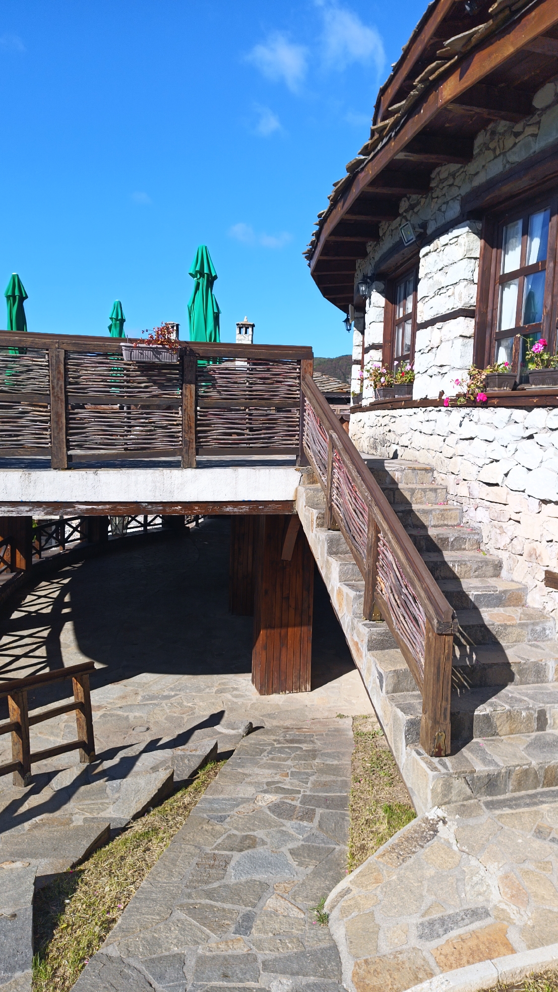 This outdoor scene, likely taken in Vrata, Bulgaria, features a stone building with wooden accents. The building has a layered, stone-tiled roof and white stone walls with dark wood trim around the windows. Flower boxes with pink flowers adorn the windowsills. A set of stone stairs with a wooden railing leads up to the building. To the left of the stairs is a raised wooden platform with a woven wooden railing. Green umbrellas are visible above the railing, suggesting an outdoor seating area. Below the platform, there is a paved area with a partial view of wooden structures. The ground is paved with irregular stone tiles, and a narrow strip of grass runs alongside the path. The sky is bright blue with a few scattered clouds. A bench is visible in the bottom left corner.