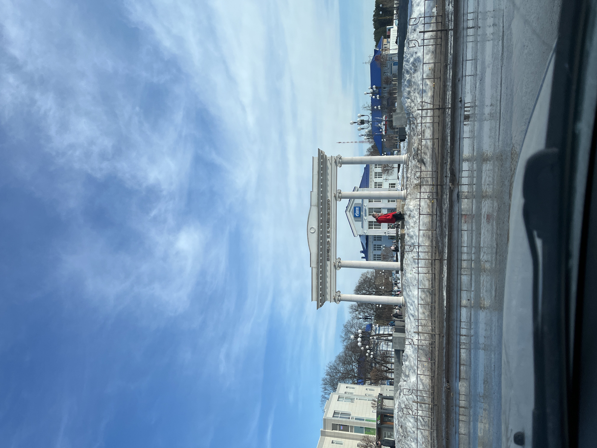 The image depicts a view from inside a vehicle, looking towards an architectural structure with classical-style columns and archways. The sky above is blue with streaks of wispy clouds.
The architectural structure appears to be a city monument or entrance to a square. The text "ПЛОЩАДЬ ЧАЙКОВСКОГО" is visible on the structure, indicating that this is located at Chaikovsky Square. A building with a blue roof and white walls is visible behind the structure.
To the right of the structure, there are metal fences, patches of snow, and wet pavement. A person wearing a red jacket is visible near the base of the monument.
The image suggests a daytime scene in the city of Chaikovsky, Russia, with a clear sky and possible melting snow. The vehicle is parked by a street, or intersection of the city.