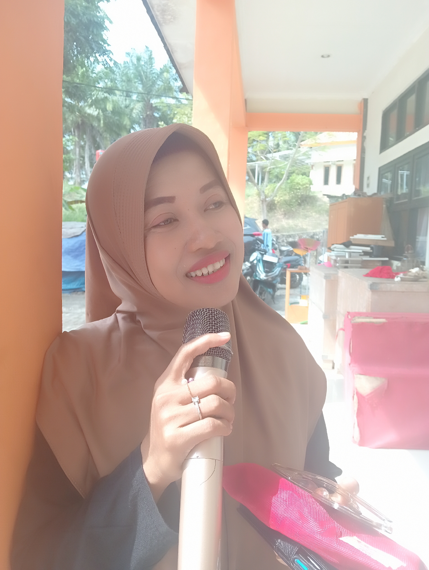 A woman wearing a tan hijab and dark clothing is holding a microphone with her right hand. She is smiling and looking to her right. Two rings are visible on her right hand: one with a square gem and another with a pearl. She is standing outdoors next to an orange pillar, with a building and trees in the background. A pink bag and a pair of clear-rimmed glasses are visible in the foreground. The lighting suggests it is daytime. The location is the city of Wanasalam, Indonesia.