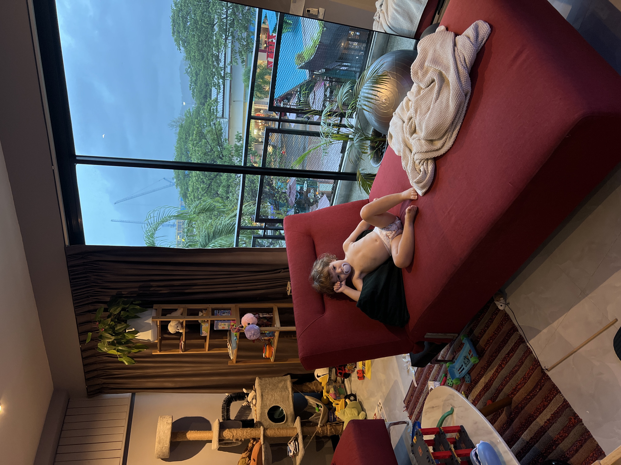 A young child, wearing only a diaper, lies on a red sofa, their legs propped up. The child has a pacifier in their mouth and appears relaxed. A light-colored blanket is draped over the sofa. The scene is indoors, with a large window revealing an outdoor view. The sky outside is overcast, suggesting it is either dawn or dusk, or possibly a stormy day. Trees and some industrial structures, possibly cranes, are visible through the window. The room appears to be a living area, with shelves holding books and toys, a cat tree, and a rug on the floor. The overall mood seems calm and domestic.