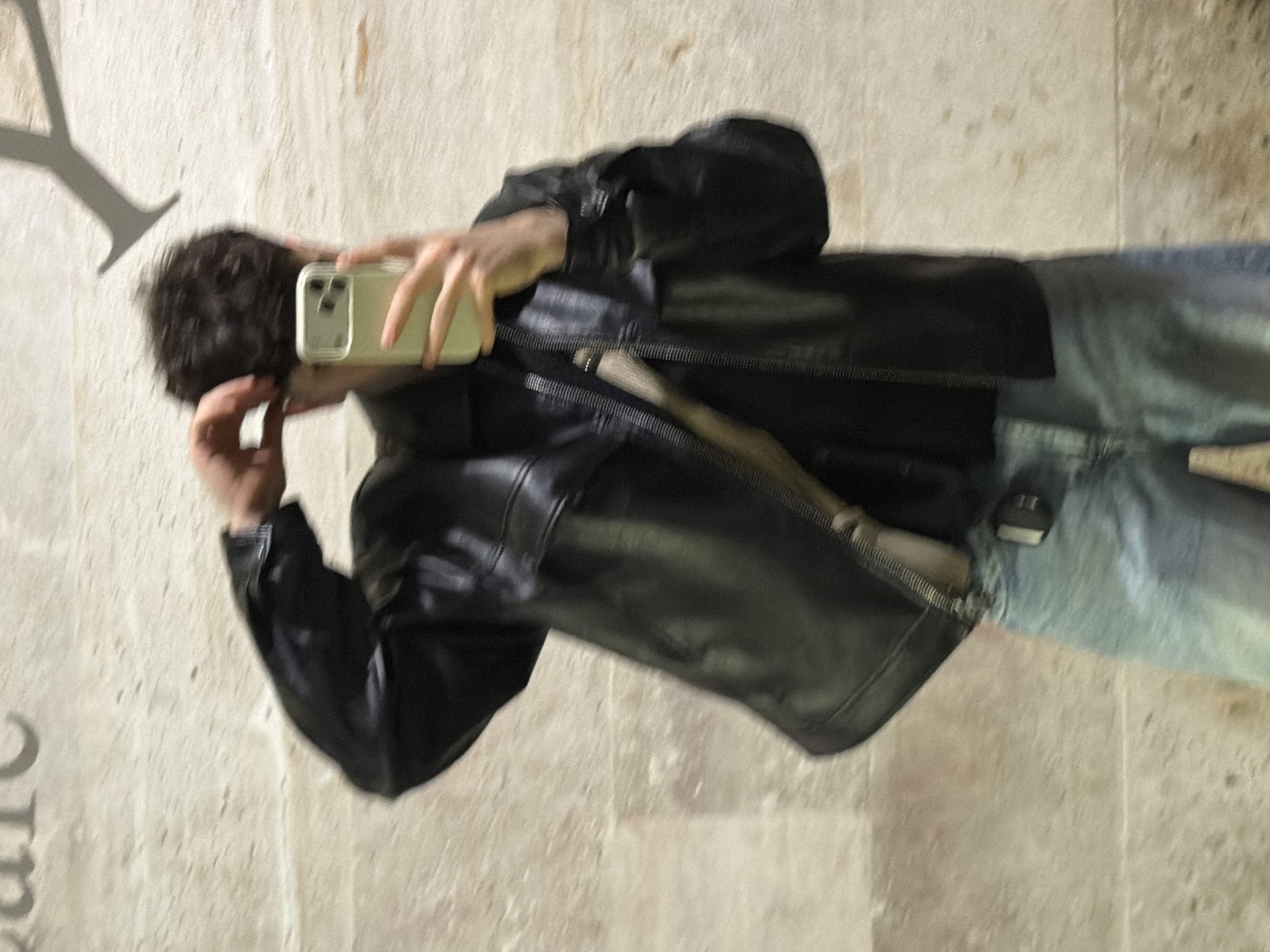 A person is taking a selfie in Tbilisi, Georgia. They are wearing a black leather jacket and blue jeans. The person is holding a smartphone with a light green case. The background appears to be a textured stone wall. There is a large, dark shape on the wall to the left of the person, resembling a letter 'Y'. The person has short dark hair.
