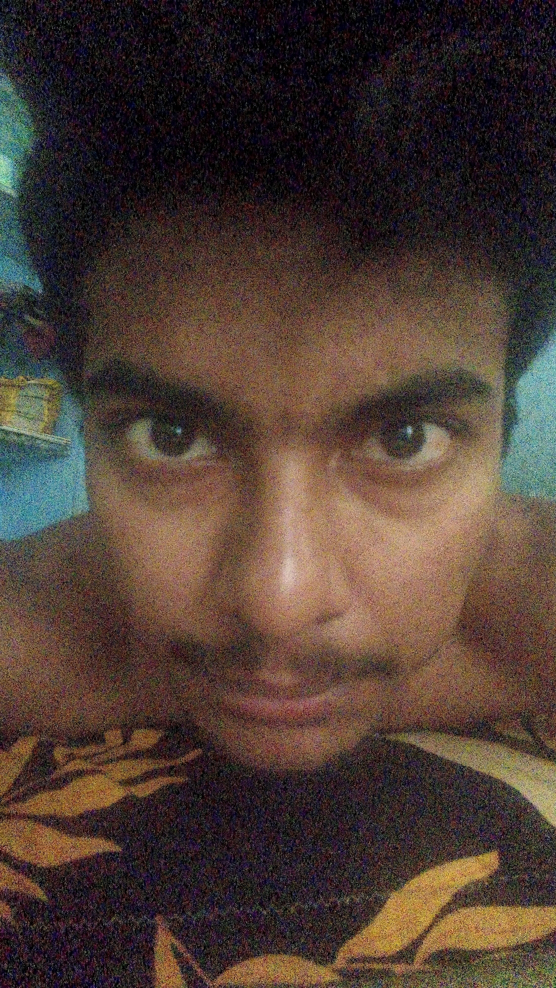 The image is a close-up, grainy selfie of a young man's face. He appears to be lying down, with his head tilted slightly back. His eyes are open and looking directly at the camera, and his mouth is closed, with a slight hint of a mustache visible above his upper lip. He has dark, curly hair and dark eyebrows. The background is blurry, but appears to be a light blue wall with some objects on a shelf to the left. The lower portion of the image shows a patterned fabric, possibly a bedspread or blanket. The overall lighting is dim, with a visible grain to the image, suggesting it was taken in low light conditions with a mobile phone camera. The setting is likely domestic, given the indoor background and bedding. There is no specific indication of the location within Karuppayurani, India, beyond it being an indoor personal space.