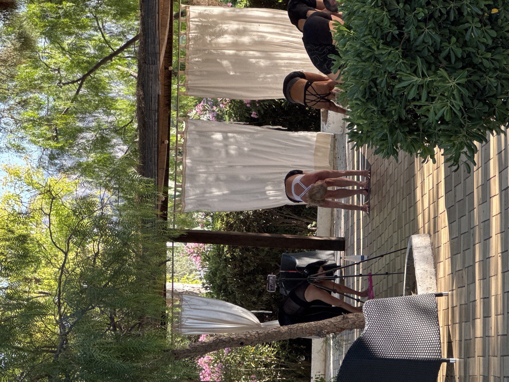 The image shows a group of women practicing yoga outdoors in Isola delle Femmine, Italy. They are performing downward-facing dog poses on a tiled patio. The background features white curtains hanging from a covered structure, lush green foliage, and pink flowers. A tripod with a smartphone is set up, presumably for recording the session. The women are dressed in athletic wear. The scene is bathed in warm sunlight, casting shadows across the patio.