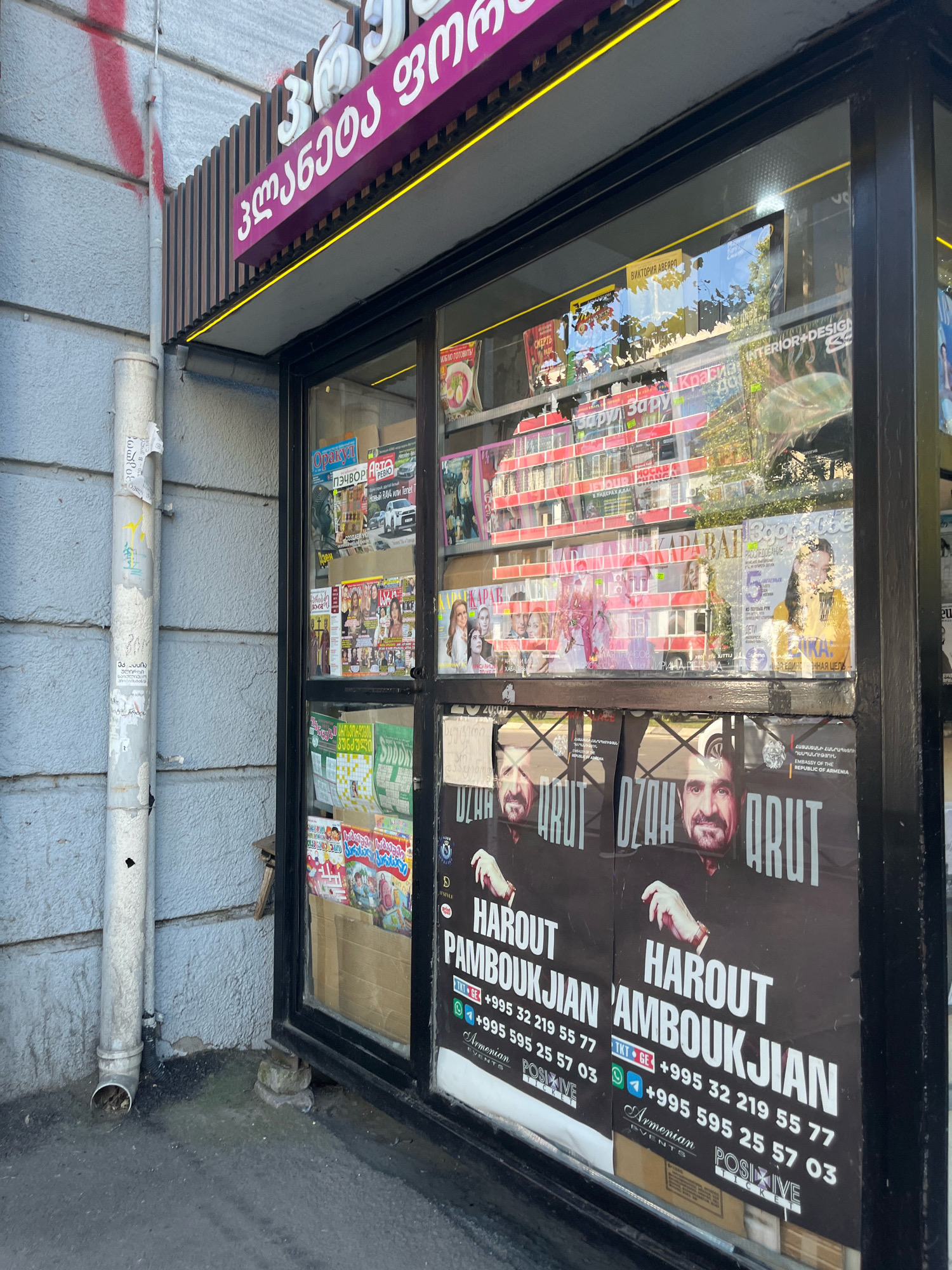 The image shows a street kiosk in Tbilisi, Georgia, with the name of the kiosk written in Georgian script. The kiosk displays various magazines and posters, with two prominent posters featuring portraits of two men. The posters also contain text in Georgian and Armenian script, along with phone numbers. The kiosk is situated on a street with a textured concrete wall to the left and a gray pavement in the foreground. A white downspout runs along the wall. The kiosk has glass windows displaying the merchandise inside. The overall scene suggests a commercial establishment offering media publications in a street setting.