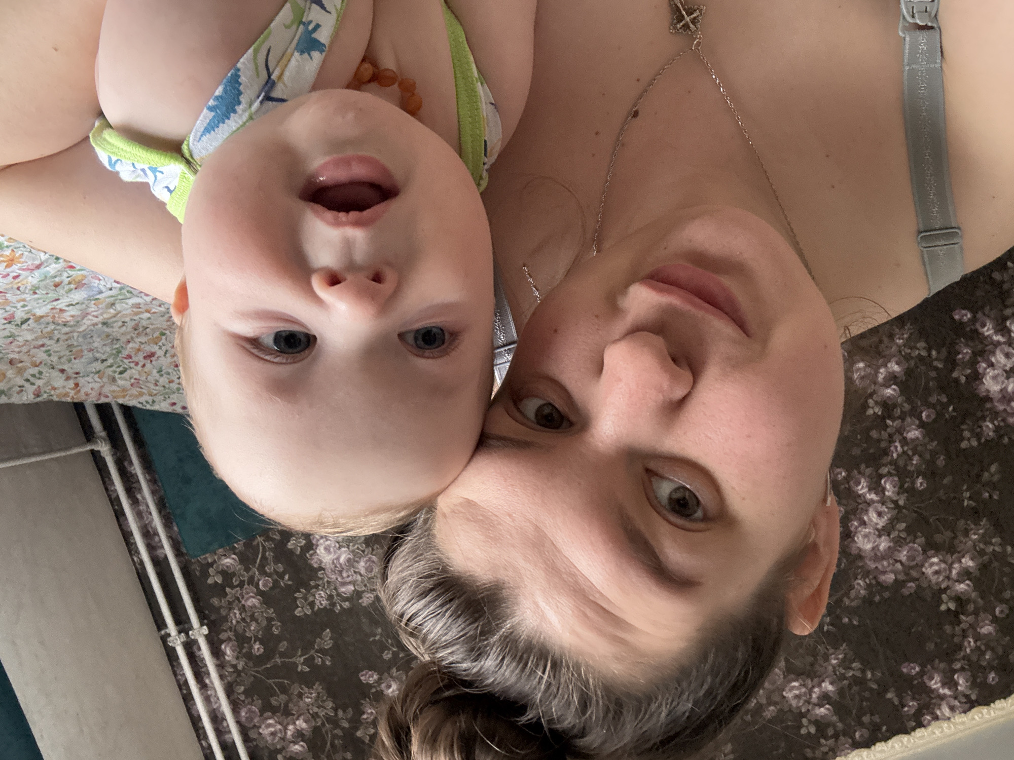 A selfie photograph features a woman and a baby. The baby, positioned above the woman, is lying on their back wearing a sleeveless top with blue and green designs. The baby's mouth is open, and they are looking at the camera. The woman is lying on her back below the baby, also looking at the camera. She is wearing a necklace with a cross pendant and a bra with a visible strap. The background consists of patterned bedding and what appears to be a wall or structure. The photo is rotated 180 degrees. There is no indication of this being in Марковский, Russia.