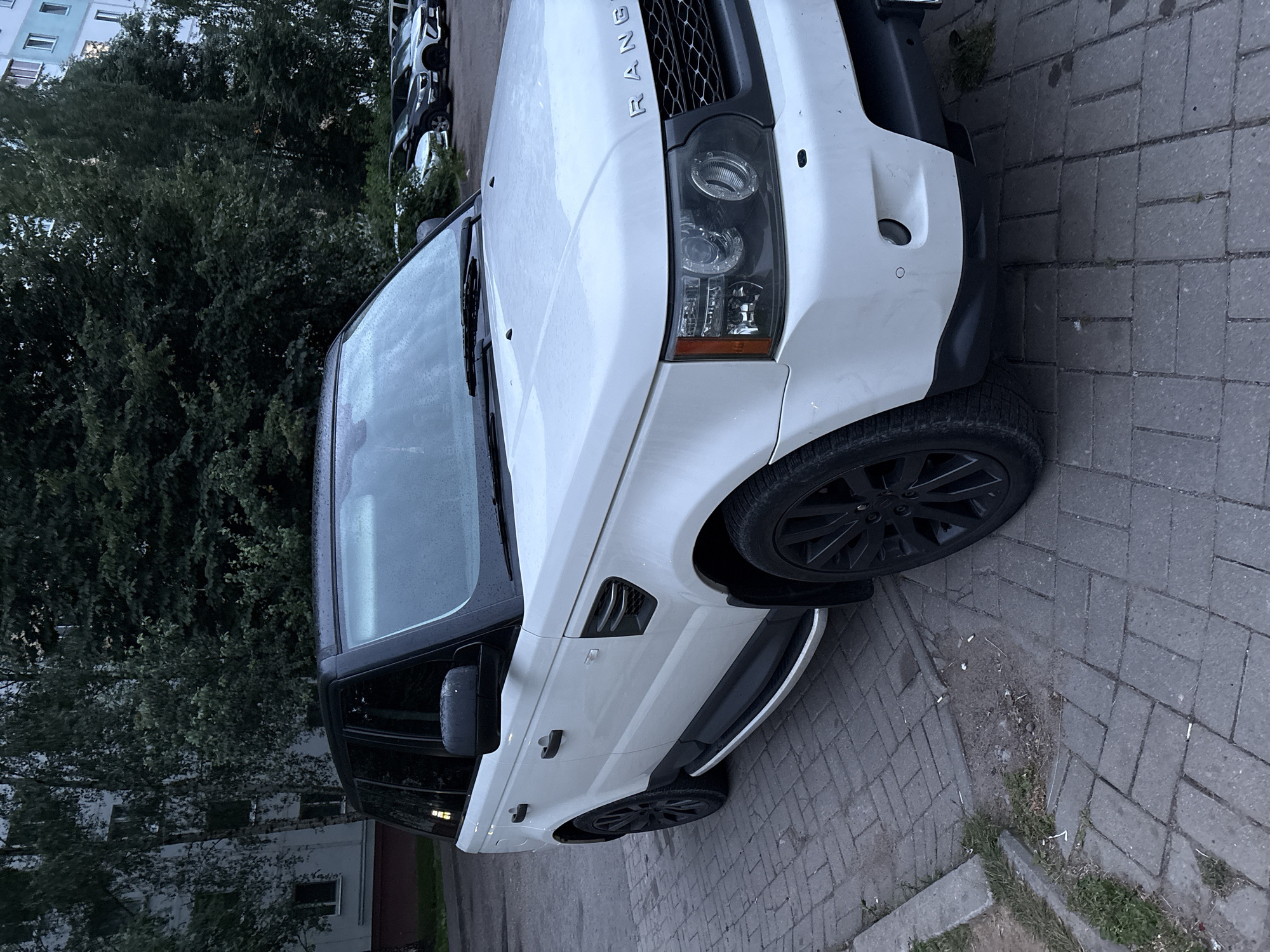 A white Range Rover Sport is parked on a paved surface in front of a building and trees. The car is facing the right side of the image. The building appears to be an apartment building with windows. The paved surface is made of grey rectangular bricks. There are trees and foliage in the background. In the distance, other cars are parked in a lot. The sky is overcast, and there are visible water droplets on the car's windshield. The scene appears to be a residential area in the city of Vitsebsk, Belarus.