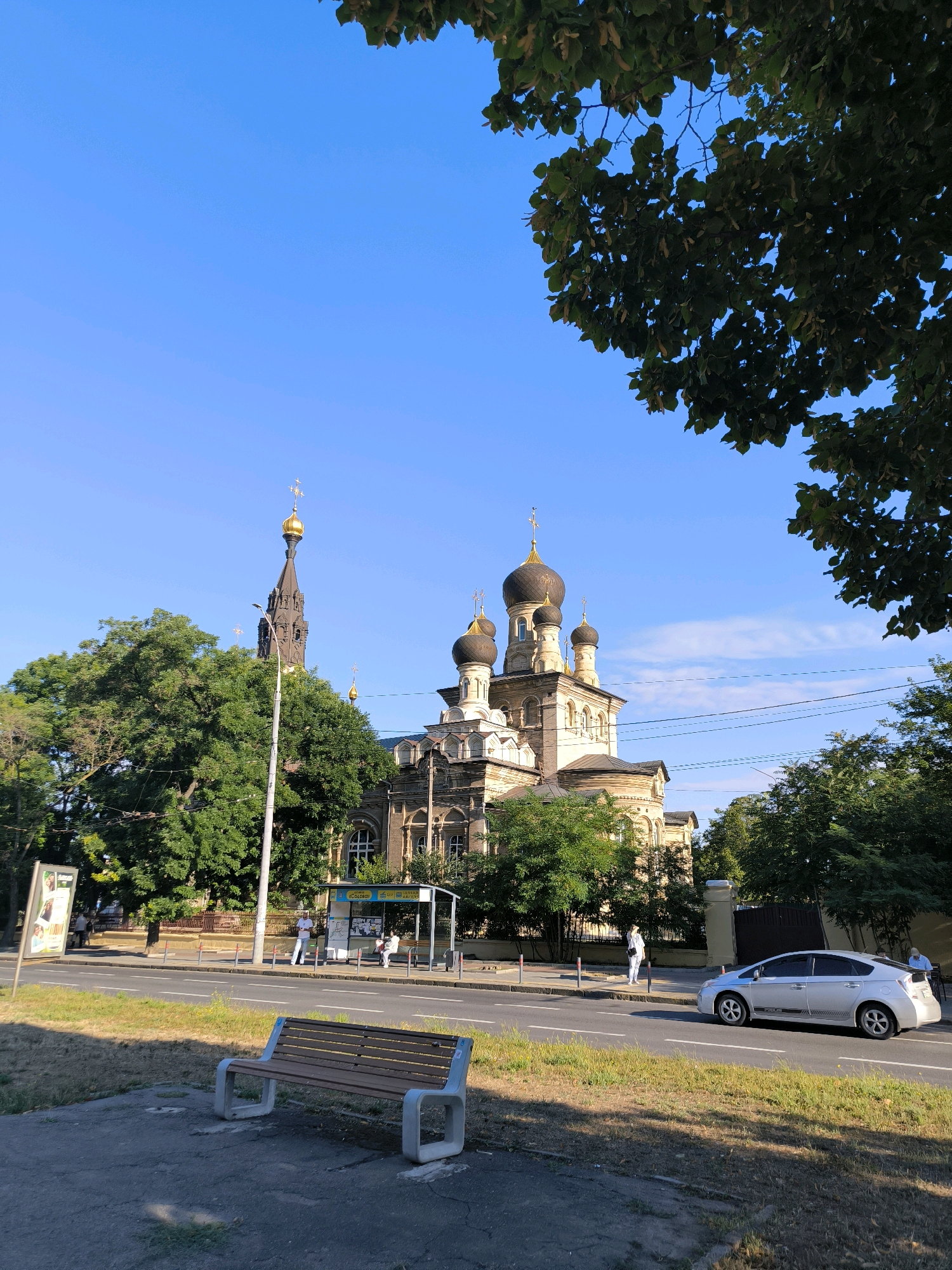 This is a ground-level, eye-level, wide shot photograph of the Transfiguration Cathedral in Mykolaiv, Ukraine. The cathedral is a large, tan building with multiple domes and a tall, slender bell tower. The cathedral is surrounded by lush green trees. In the foreground, there is a sidewalk with a park bench and a paved area. A street runs parallel to the sidewalk, with a silver Toyota Prius driving on it. There is a bus stop on the side of the street, with a few people waiting. The sky is a clear blue with a few wispy clouds. The sun is shining brightly, casting shadows across the scene.