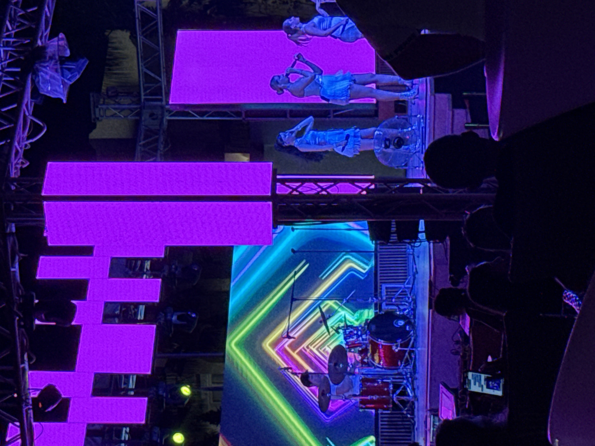 This image captures a vibrant outdoor evening concert scene in Sharm El Sheikh, Egypt. The stage is brightly lit with large purple LED screens forming a backdrop, and a dynamic, colorful geometric light display is projected behind a drummer. Three female singers, dressed in light blue, stand on a raised platform on the right side of the stage, holding microphones and performing. In the foreground, the silhouettes of audience members are visible, seated and observing the performance. The overall atmosphere is energetic and festive, illuminated by the colorful stage lights against the night sky.
