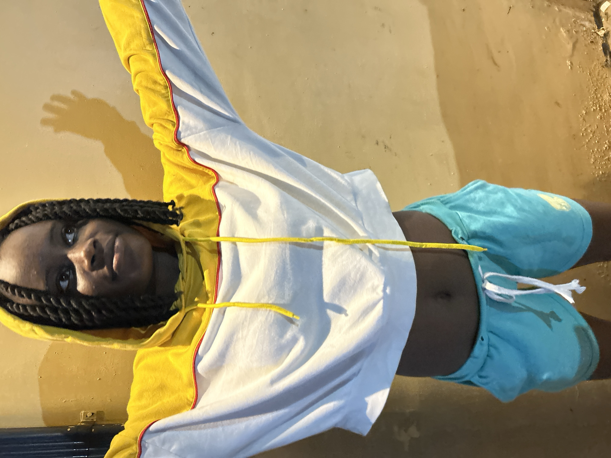 A young person with dark skin and braided hair stands with arms outstretched against a textured, yellowish-brown wall. The person wears a cropped white hooded sweatshirt with a yellow yoke and red trim, and light blue athletic shorts. Yellow drawstrings hang from the hood of the sweatshirt. A shadow of the person's outstretched hand is visible on the wall behind them. The scene appears to be outdoors, possibly in a courtyard or alleyway in Ihiagwa, Nigeria, given the wall texture and lighting.