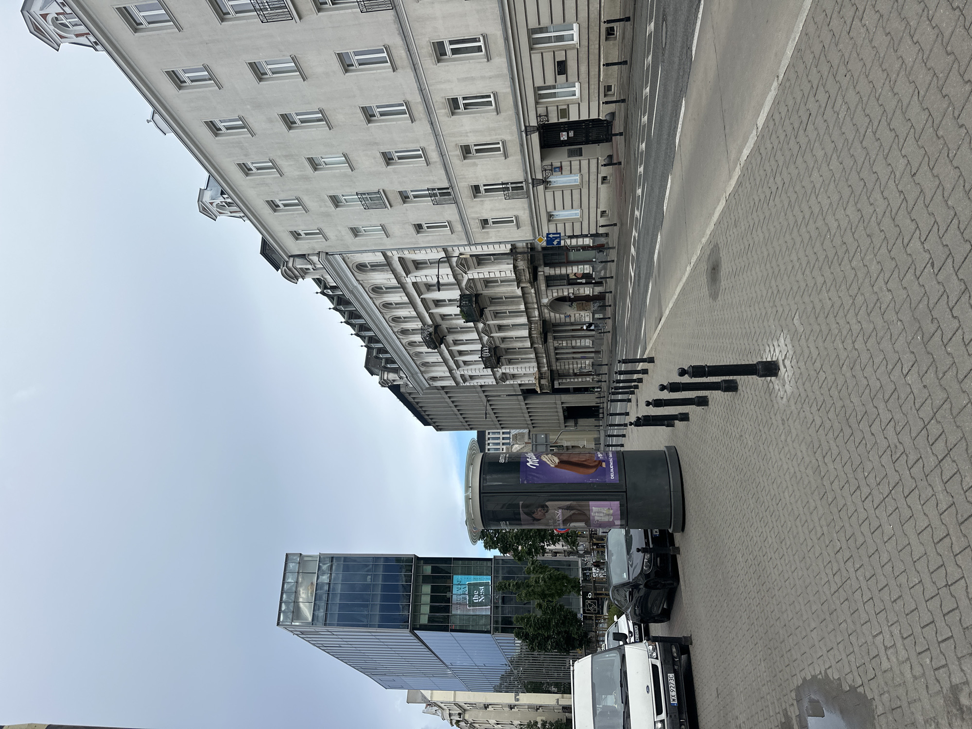 This is a street view in Warsaw, Poland, showing an urban environment with a mix of modern and older architecture. On the left, a contemporary glass-fronted building with multiple stories stands prominently. Adjacent to it is a cylindrical advertising column displaying posters. To the right of the column, a black car is parked, and further to the right, a white van is also visible.
The sidewalk is paved with interlocking gray bricks. A street stretches across the middle ground, with visible lane markings. Along the right side of the street, a row of pale, multi-story buildings with traditional architectural elements, including mansard roofs and balconies, lines the pavement. These older buildings contrast with the modern glass structure on the left. The sky above is overcast.