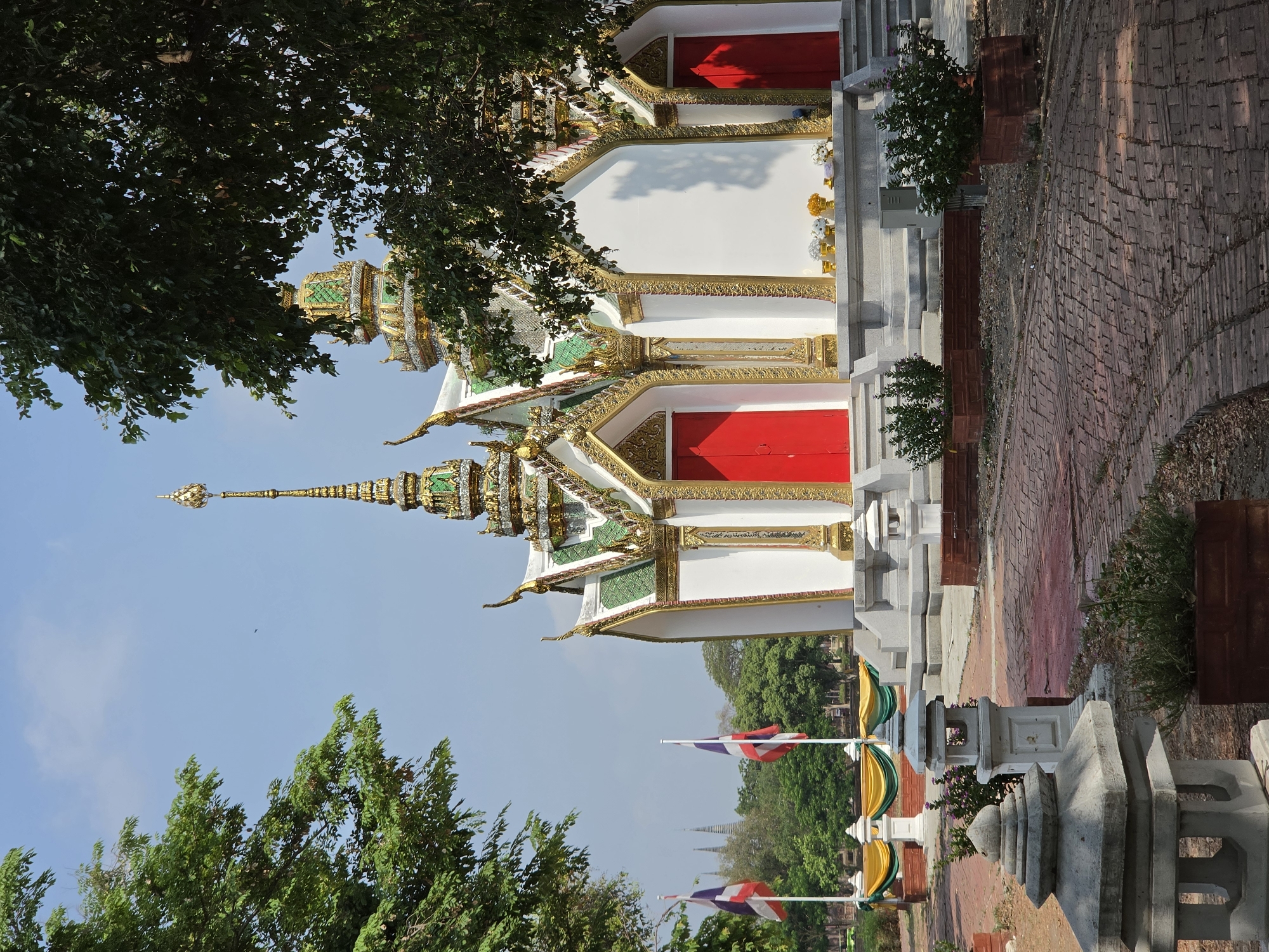 The image shows a temple in Phra Nakhon Si Ayutthaya City Municipality, Thailand. The temple building is white with intricate gold and green detailing. It has a tall, ornate spire. There are red doors on the side of the temple. Thai flags are visible in the background. A brick walkway and stone structures are in the foreground. Trees partially obscure the view of the temple. The sky is light blue.