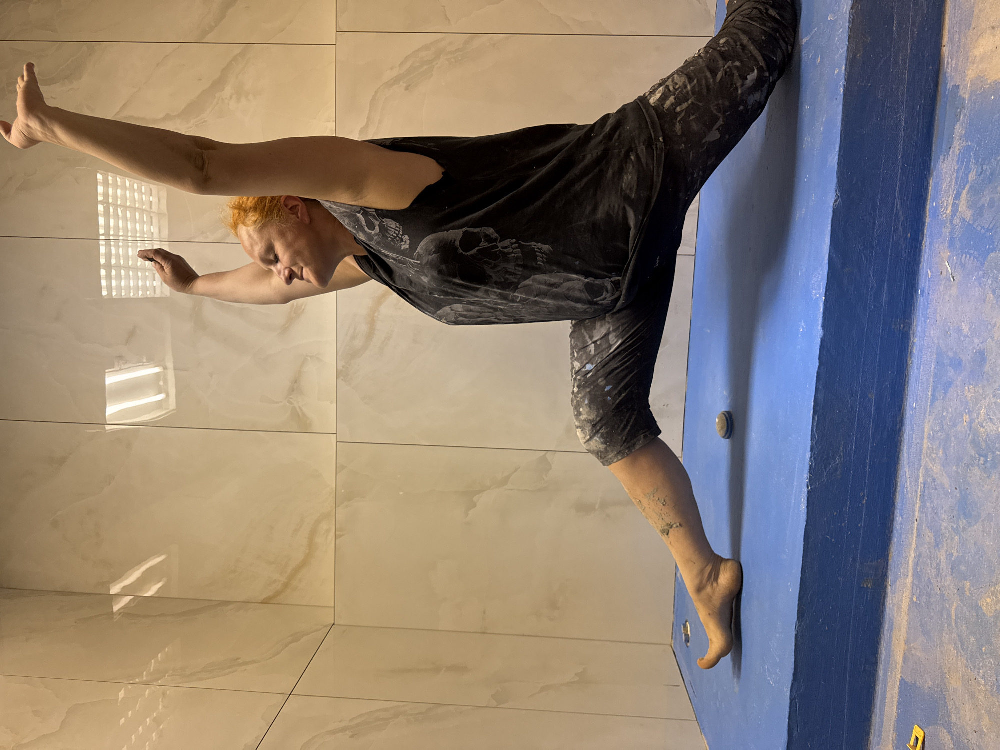 A woman is shown mid-exercise in what appears to be an indoor setting with tile walls and a blue climbing wall. The woman has reddish-orange hair and is wearing a black tank top with skull imagery and black pants with a speckled pattern. She is in a standing split pose, with her left hand on the tile wall and her right hand extended upward. Her right foot is touching the climbing wall. The environment is a room with glossy tile walls that reflect light, suggesting it could be a modern interior space. The location is Chaikovsky, Russia.