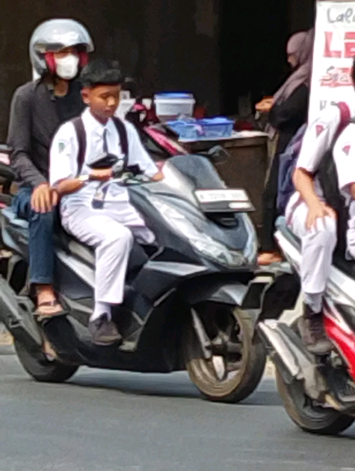 In Manaruwi, Indonesia, two individuals are riding a black motorcycle. The person in the front, a young male wearing a white school uniform with a dark tie and black shoes, is seated. The person behind, the driver, is wearing a dark jacket, blue jeans, and flip-flops, with a silver helmet and a white mask. A sign with red and white lettering is visible to the right of the scene, along with a woman wearing a headscarf. The motorcycle is on a paved road.