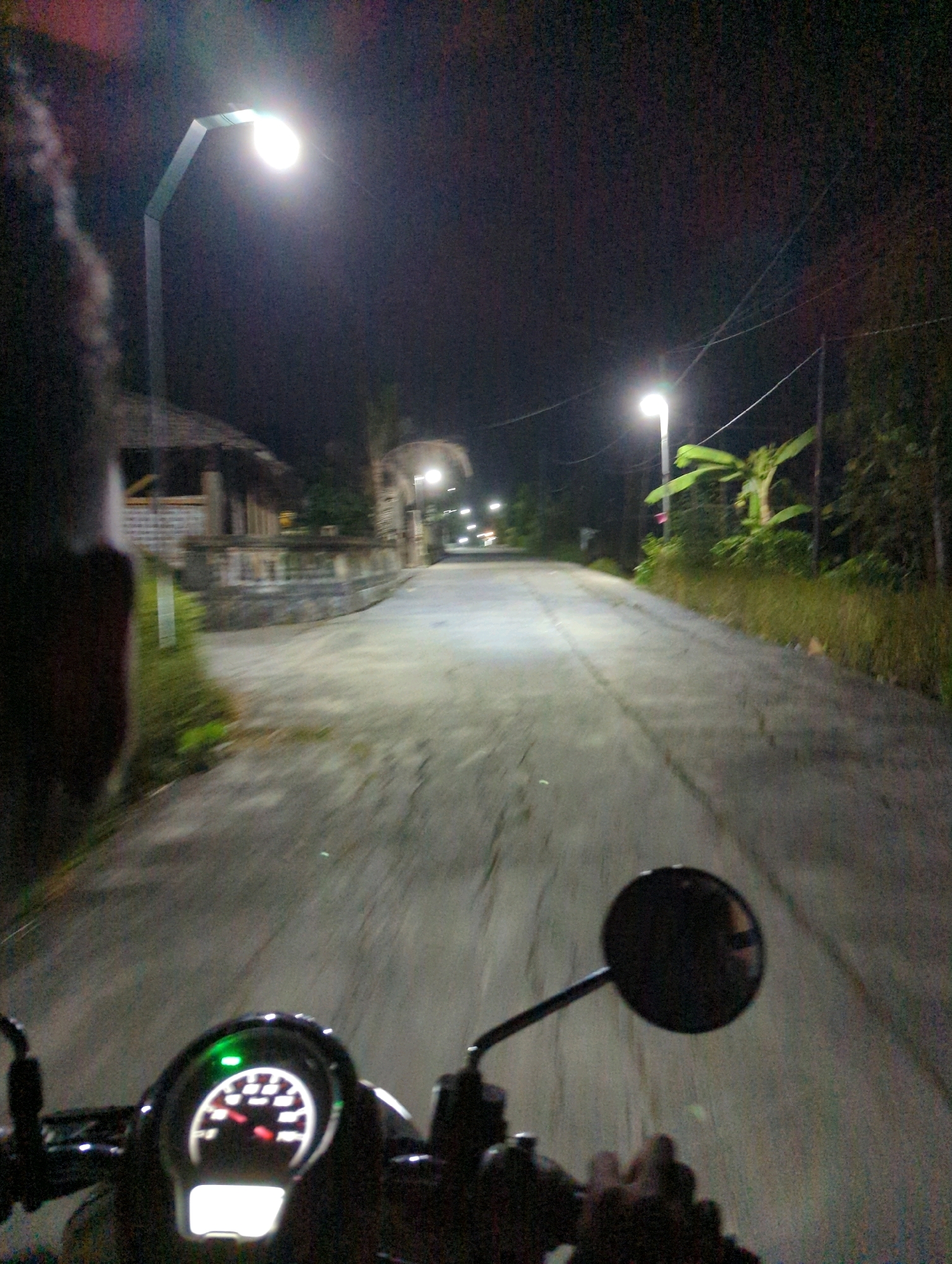 The media file captures a first-person perspective of someone riding a scooter at night in Kunti, Indonesia. The scene is illuminated by streetlights, casting a glow on the paved road ahead. In the foreground, the scooter's dashboard is visible, with a speedometer showing the speed and a green indicator light illuminated. A side mirror is also present, reflecting the road.
To the left of the road, there's a glimpse of a building with a thatched roof and what appears to be a stone wall, suggesting a traditional or rustic setting. Further down the road, another streetlight is visible, along with distant lights that could indicate other vehicles or buildings. The surrounding area is dark, with trees and vegetation lining the right side of the road, typical of a rural or suburban environment. The overall impression is of a calm, solitary night ride through a well-lit, yet quiet, part of Kunti. No text is discernible in the media file.