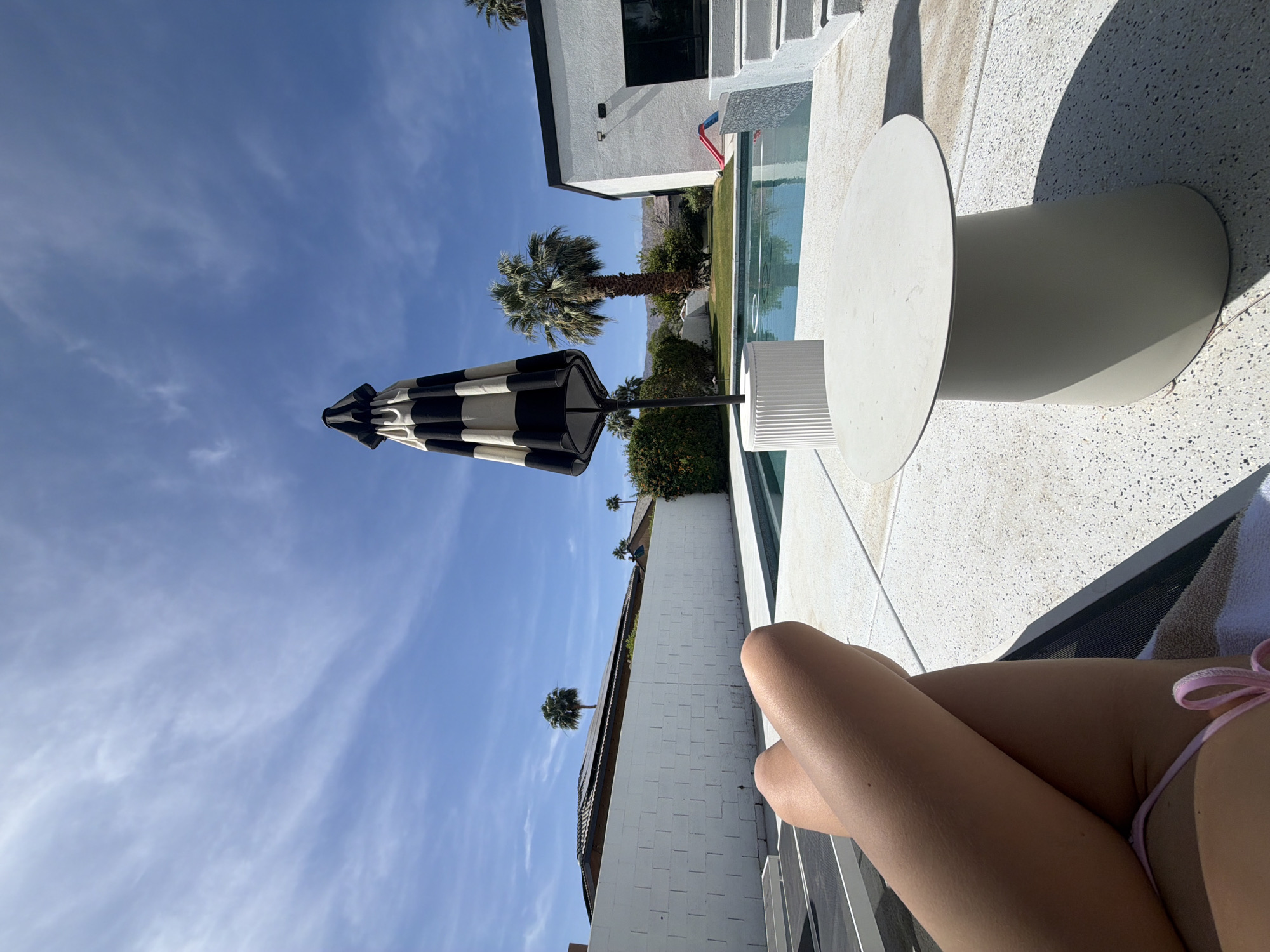 The image shows a poolside scene in Palm Desert, California, USA. The view is from a low angle, possibly from a lounge chair occupied by a person whose legs are visible in the foreground, wearing a pink bikini bottom. Adjacent to the chair is a round, white side table and beyond the table, a swimming pool with clear water. The pool is bordered by white coping and a modern white building with large windows and a red element is visible nearby. A black and white striped umbrella is set up to provide shade. Palm trees are present, contributing to the desert environment. The sky is blue with scattered clouds.