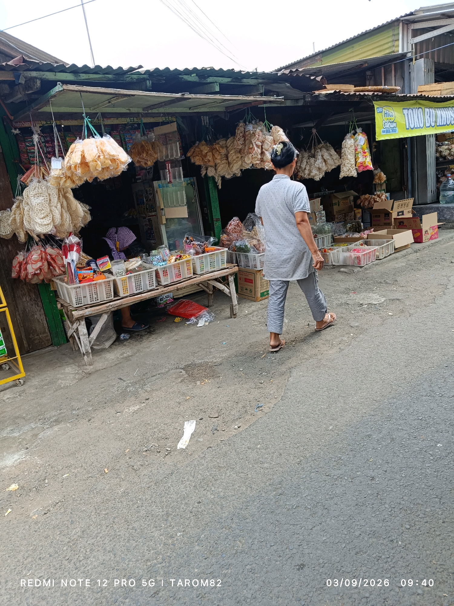 The image depicts an outdoor marketplace scene in Kandangan, Indonesia. A person wearing a grey t-shirt and striped grey pants is walking away from the viewer towards a market stall. The stall has a corrugated metal roof and is lined with various goods, including bags of snacks, crackers, and other food items displayed in white plastic baskets. A wooden table supported by stilts holds more baskets of goods. In the background, there are stacks of cardboard boxes and a large blue water jug. A yellow cart is partially visible on the left side of the image. The ground is paved with asphalt and shows signs of wear and tear. A banner with Indonesian text, including "TOKO BU MIS", is hanging from the stall. The lighting suggests it is daytime, likely morning given the timestamp at the bottom.