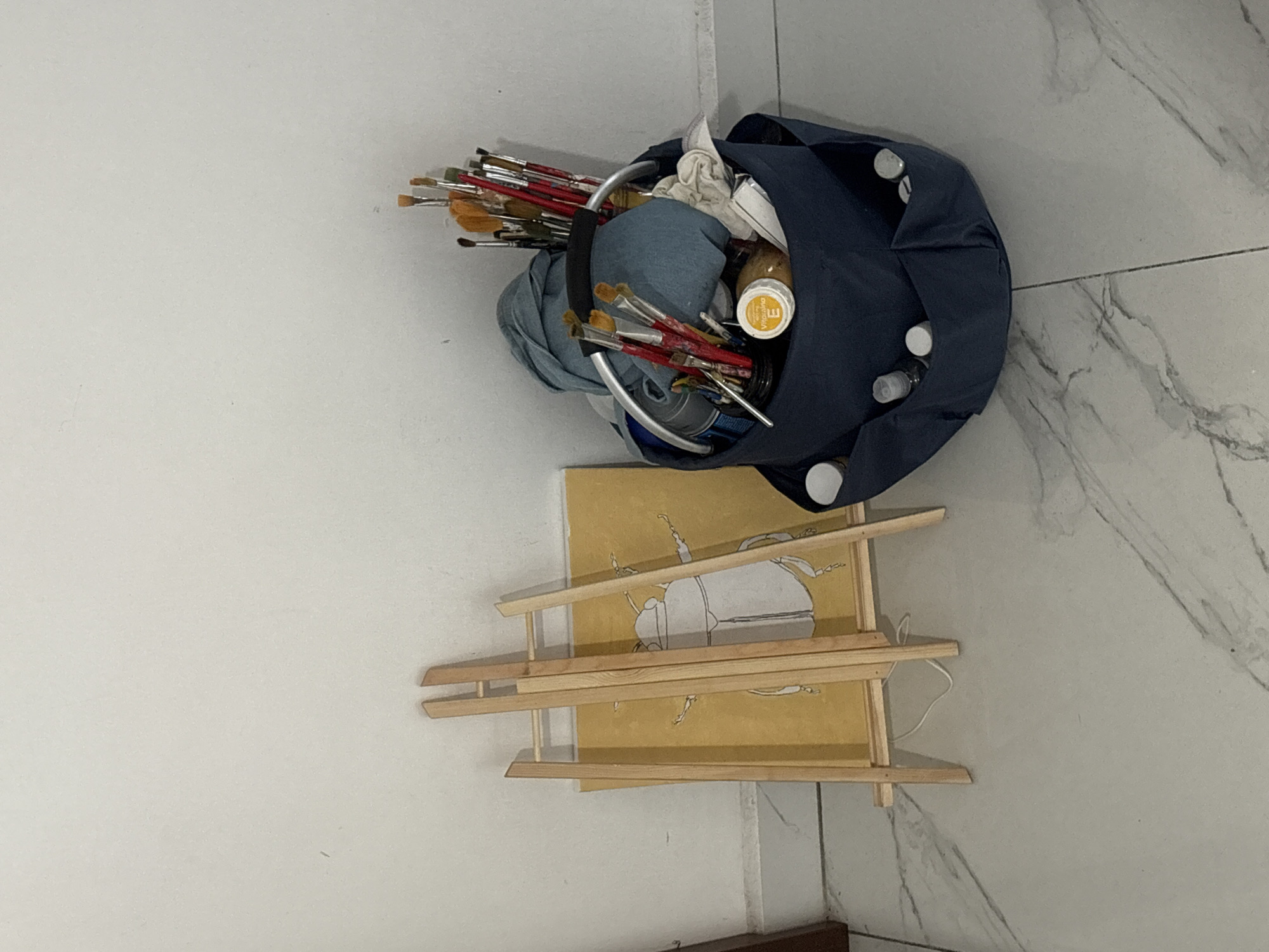 A photograph taken indoors in a room with tile flooring and off-white walls shows a collection of art supplies. A dark blue bag contains numerous paintbrushes, some with red handles and brown brush heads, a jar with a yellow label, and other items. Next to the bag, there are stacked wooden frames and a yellow canvas with a black and white drawing of a scarab beetle on it. The items are positioned on the floor, and the scene appears to be somewhat disorganized. There is no information available to determine the specific location as San Bernardino Tlaxcalancingo, Mexico. There are no people in the image.
