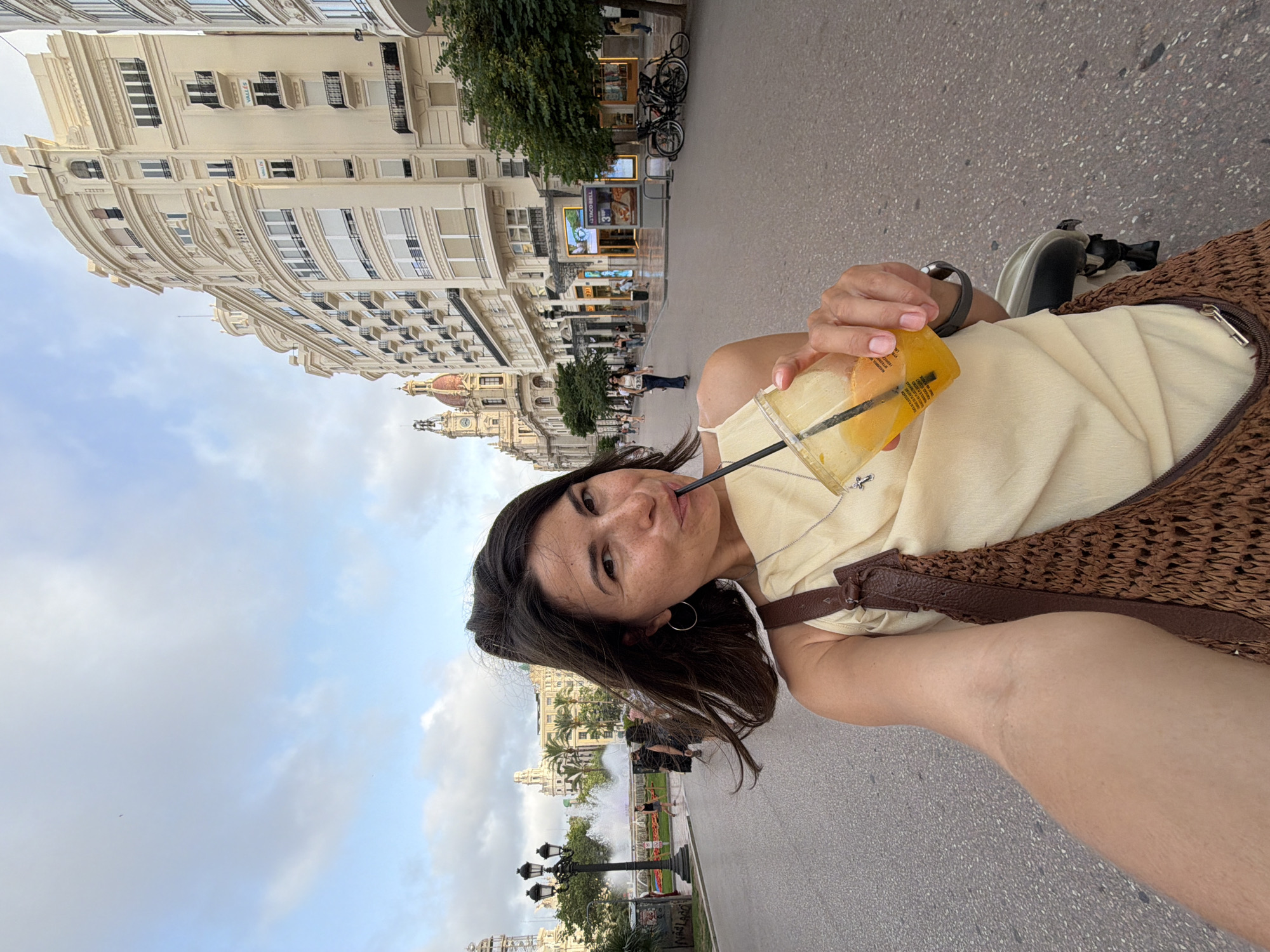 A woman, seen from the chest up, is taking a selfie in a European city. She has dark hair and is wearing a light-colored sleeveless top, a brown woven bag over her shoulder, and a necklace with a cross pendant. She is holding a clear plastic cup filled with an orange beverage and a straw, and she is sipping from it. Her left arm is visible, showing some stubble. In the background, a street stretches into the distance, lined with ornate, cream-colored buildings with balconies. There are lampposts and trees along the sidewalk. Further down the street, a fountain with colorful lights is visible, and several people are walking. Bicycles are parked on the sidewalk. The sky is cloudy.