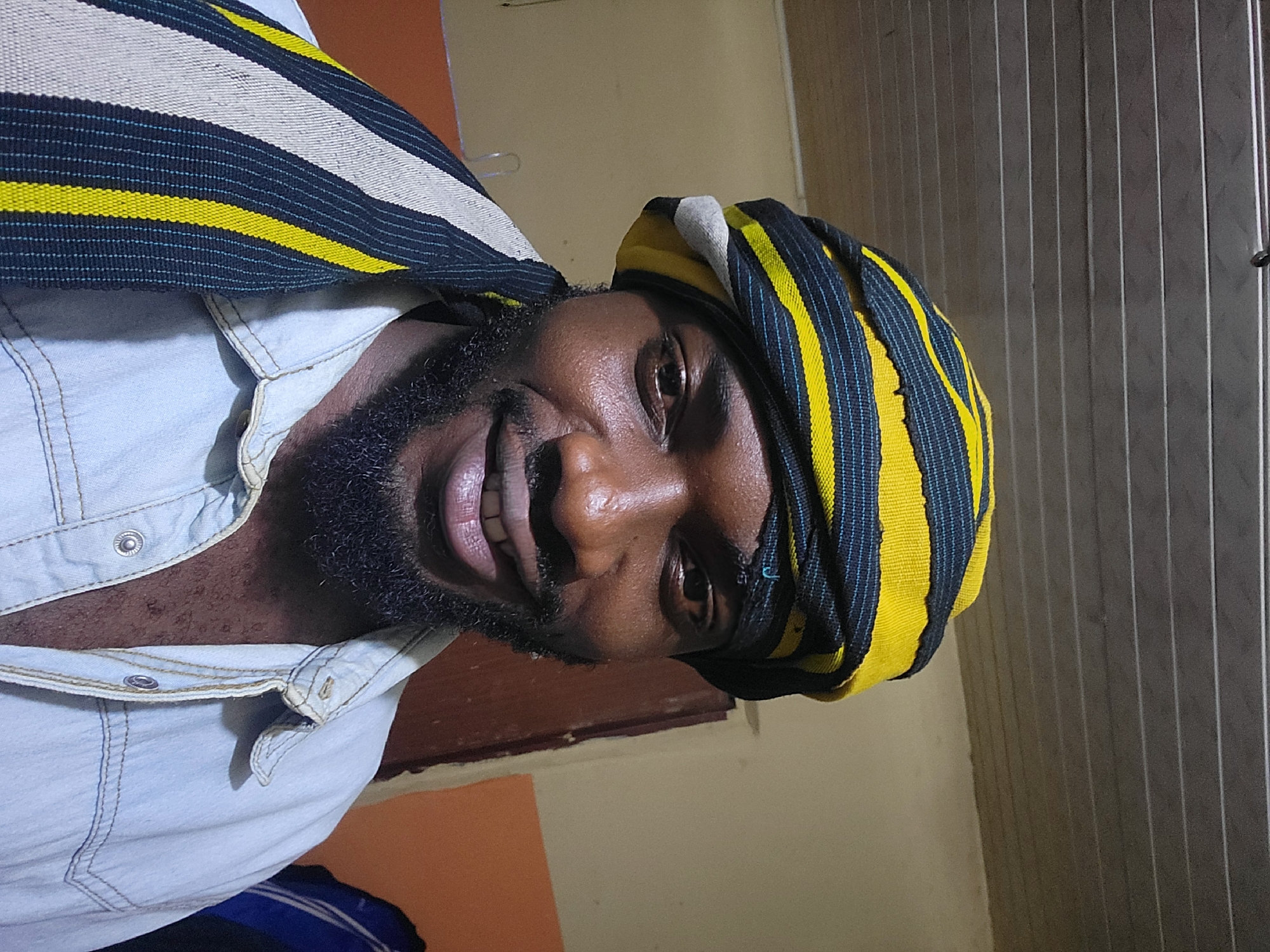 A man with dark skin and a full beard is captured in a close-up, chest-up shot, looking directly at the camera with a wide, cheerful smile that reveals his teeth. He is wearing a light blue denim shirt with visible stitching and snap buttons. Around his head, he has a distinctive headwrap made of patterned fabric featuring bold yellow and dark blue stripes, with fine light blue threads woven into the dark sections. A similar piece of striped fabric, also incorporating white or cream stripes, is draped over his left shoulder.
The man's expression conveys happiness or amusement as he poses for the photograph. The setting appears to be indoors. The background features a plain, earthy orange-brown wall on the lower left, transitioning to a lighter cream-colored wall, and then to a section of vertically striped dark grey paneling or blinds on the right. The lighting suggests an indoor environment, possibly artificial. No text is visible. This scene is located in Jos, Nigeria.