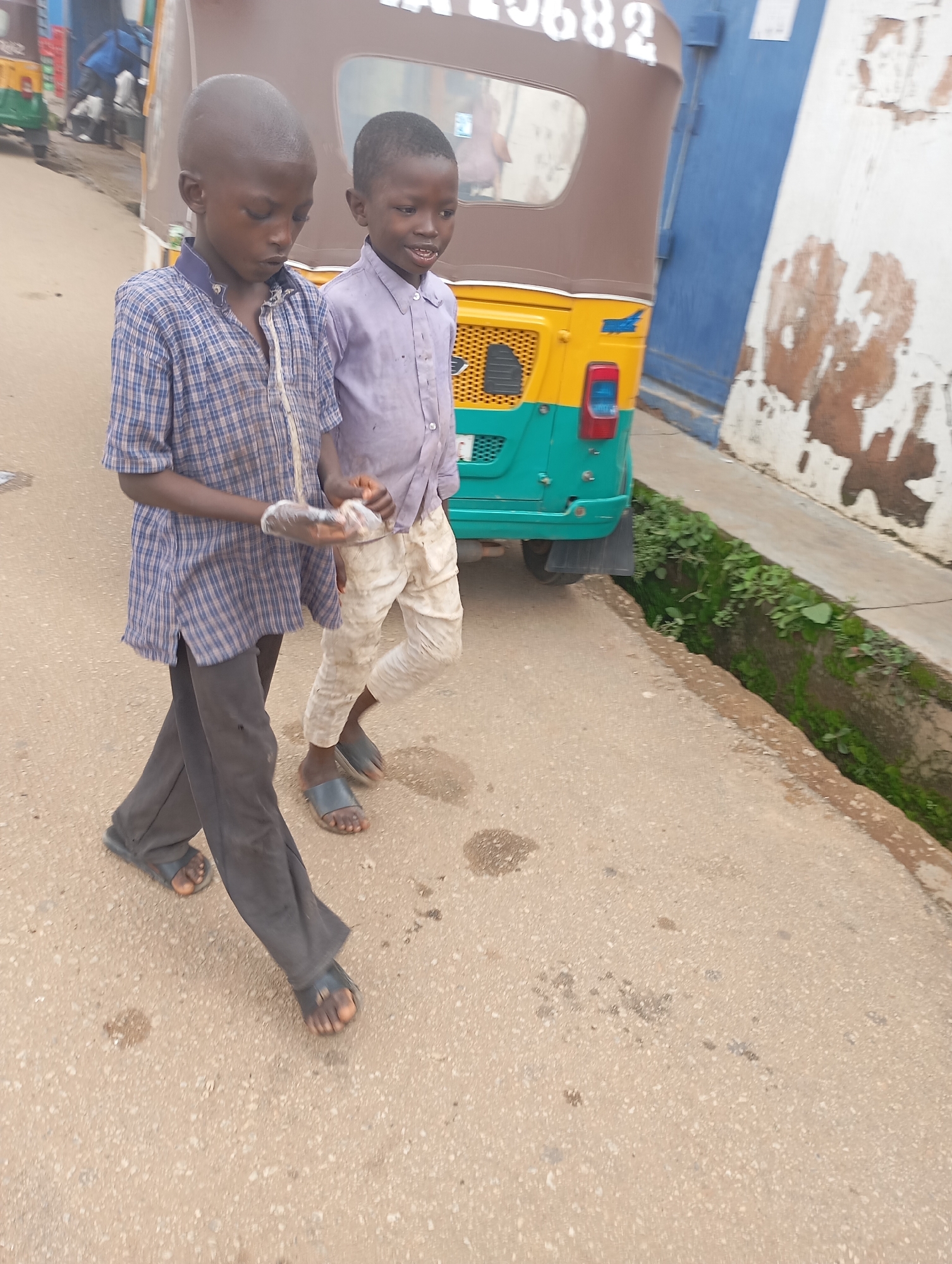 In Jos, Nigeria, two young boys walk side-by-side on a paved street. The boy on the left wears a blue and white checkered shirt and dark pants, with black sandals. He holds a clear plastic bag in his right hand. The boy on the right is dressed in a light purple shirt and light-colored pants, also wearing black sandals. They are walking towards the viewer's left. In the background, a yellow and green auto-rickshaw is parked, with a partial view of a blue wall and a white building with peeling paint. Green vegetation grows along a ditch to the right of the street.